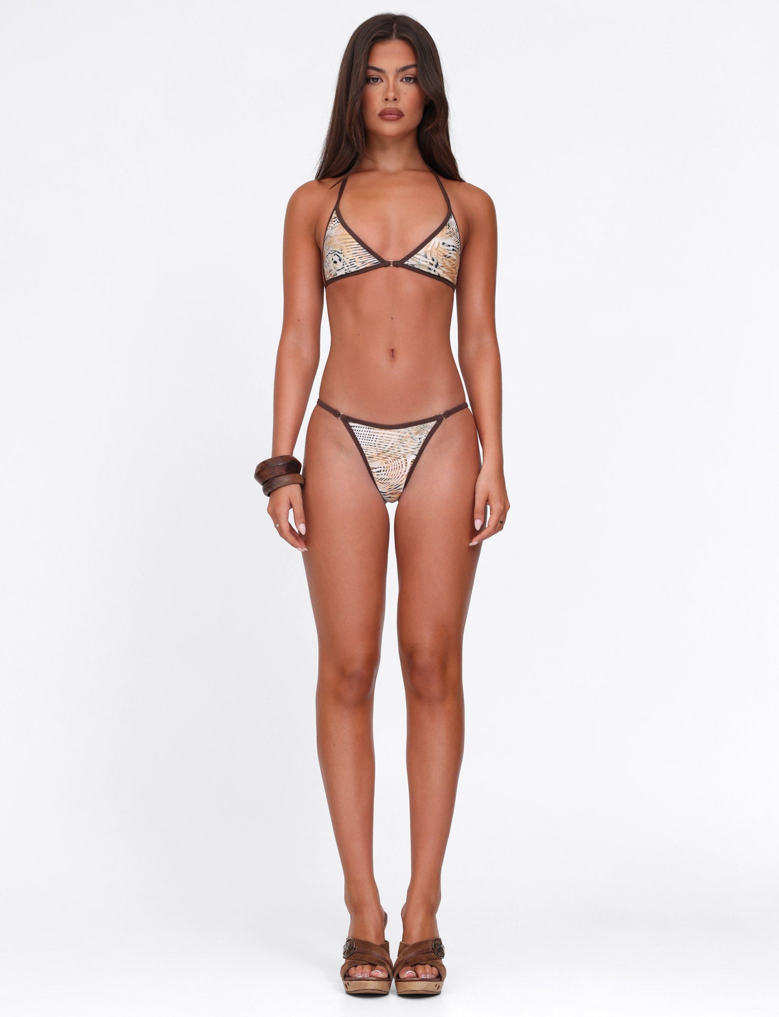 A woman with long brown hair stands facing forward in the TIGERMIST DIANA BIKINI TOP - NEUTRAL : ANIMAL : ANIMAL MARBLE, paired with brown heels and bracelets, against a plain white background. She has a neutral expression.