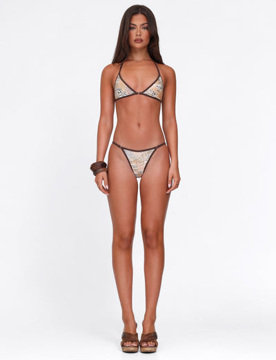 A woman with long brown hair and a neutral expression stands facing forward against a plain white background, wearing the TIGERMIST DIANA BIKINI BOTTOM in Animal Marble print - Neutral: Animal. She pairs it with brown sandals and a large brown bracelet.