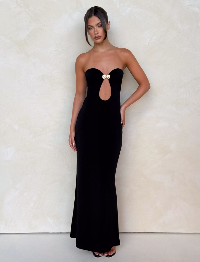 Against a light textured wall, a woman wears the TIGERMIST CECILY MAXI DRESS in black—a strapless maxi with sweetheart neckline, front keyhole cutout, and gold bust detailing. Her hair is up and she pairs it with black open-toe heels.