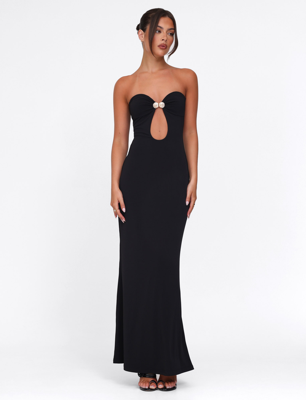A woman stands against a white background wearing the TIGERMIST CECILY MAXI DRESS - BLACK, a strapless, floor-length gown featuring a front cut-out, sweetheart neckline, and two decorative buttons at the bust. She styles her hair up and wears black heels.