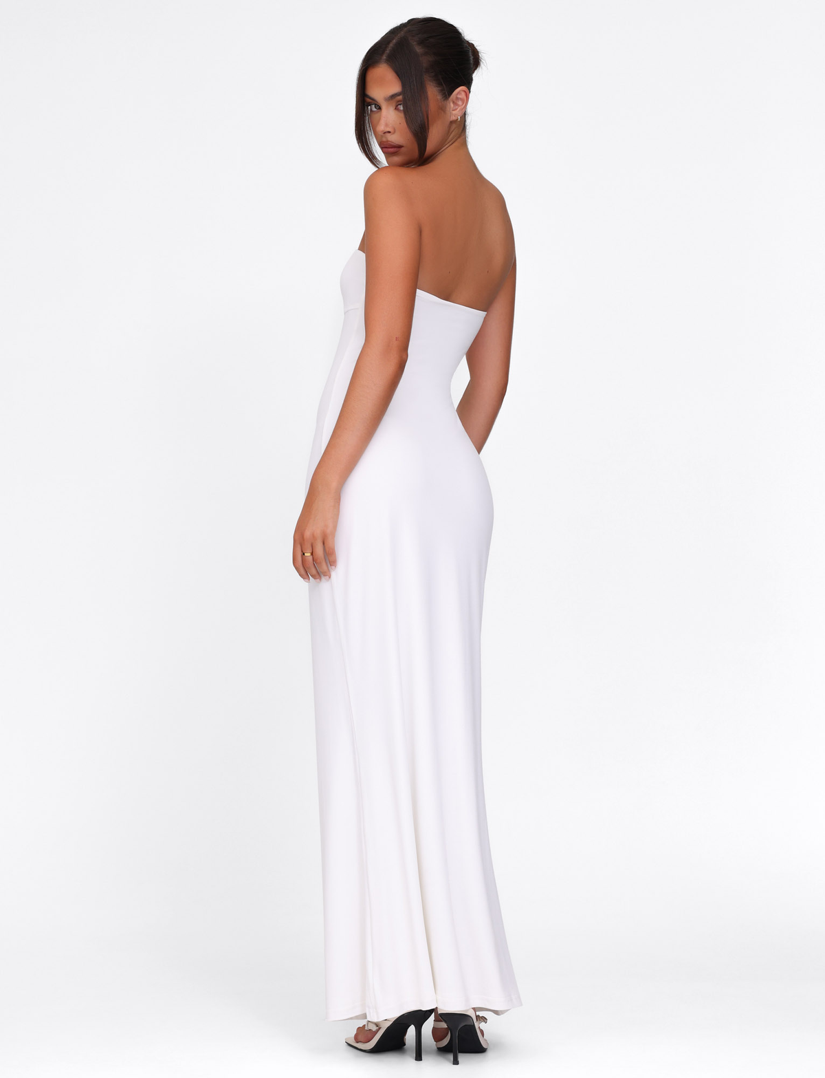 A woman poses against a white background in the TIGERMIST CECILY MAXI DRESS - WHITE, a strapless maxi with a sweetheart neckline, paired with black heels. Her hair is styled in a loose updo as she looks over her shoulder.