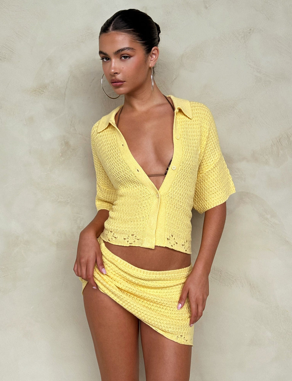 A person standing against a textured, neutral-colored wall wears the JANAI TOP - YELLOW : LEMON from TIGERMIST, consisting of a partially unbuttoned short-sleeve top and a matching mini skirt. They have hoop earrings and long hair pulled back. The pose is relaxed and confident.