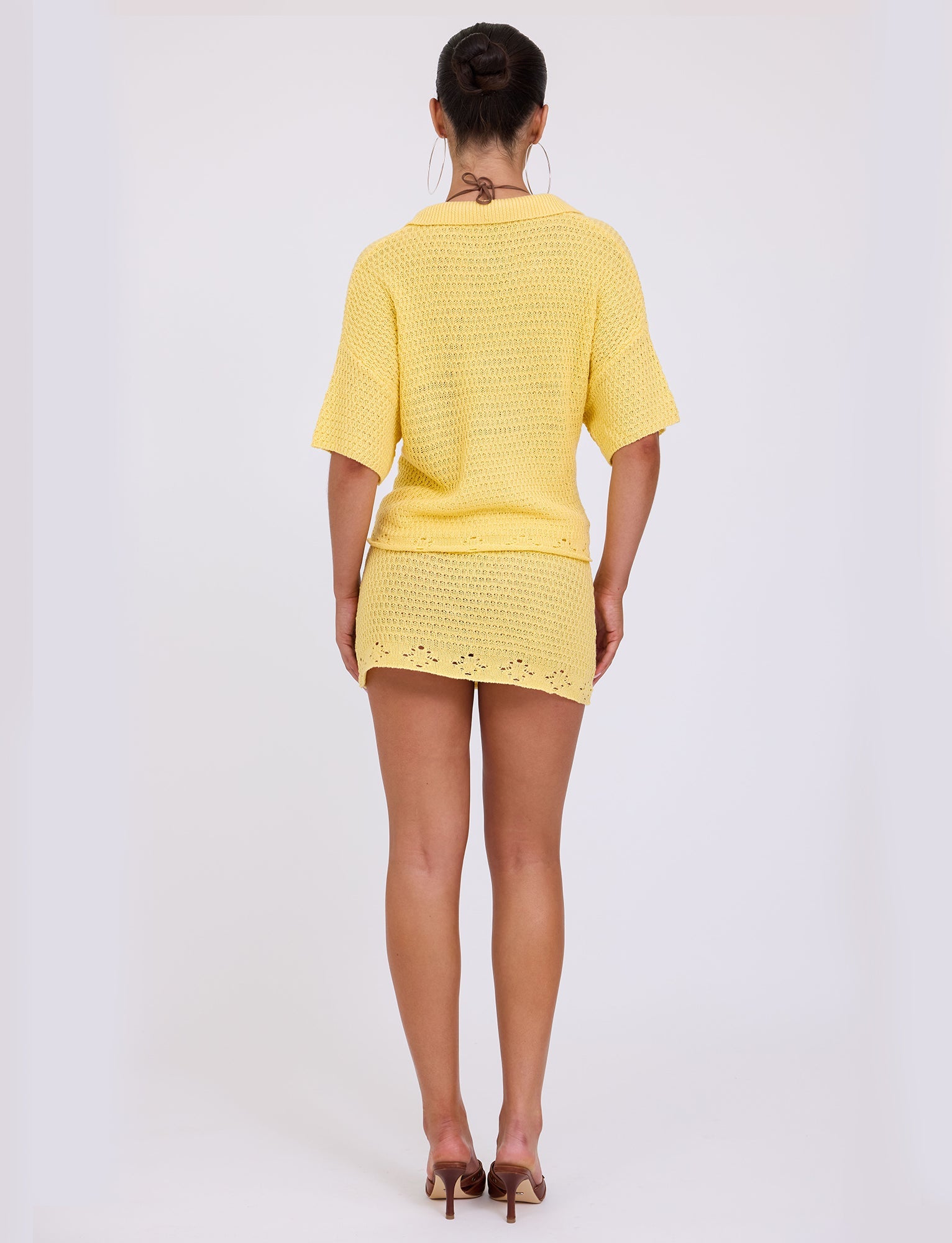 A person with their hair in a bun is standing and facing away from the camera. They are wearing a yellow knit top and the JANAI SKIRT - YELLOW : LEMON from TIGERMIST, paired with brown high-heeled shoes. The background is plain white.