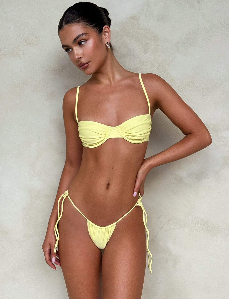 A woman in a yellow Tigermist Khessa Bikini Bottom - Lemon poses in front of a neutral, textured background. She has her hair pulled back and is looking to the side, showcasing the two-piece swimsuit with spaghetti straps and side-tie bottoms.