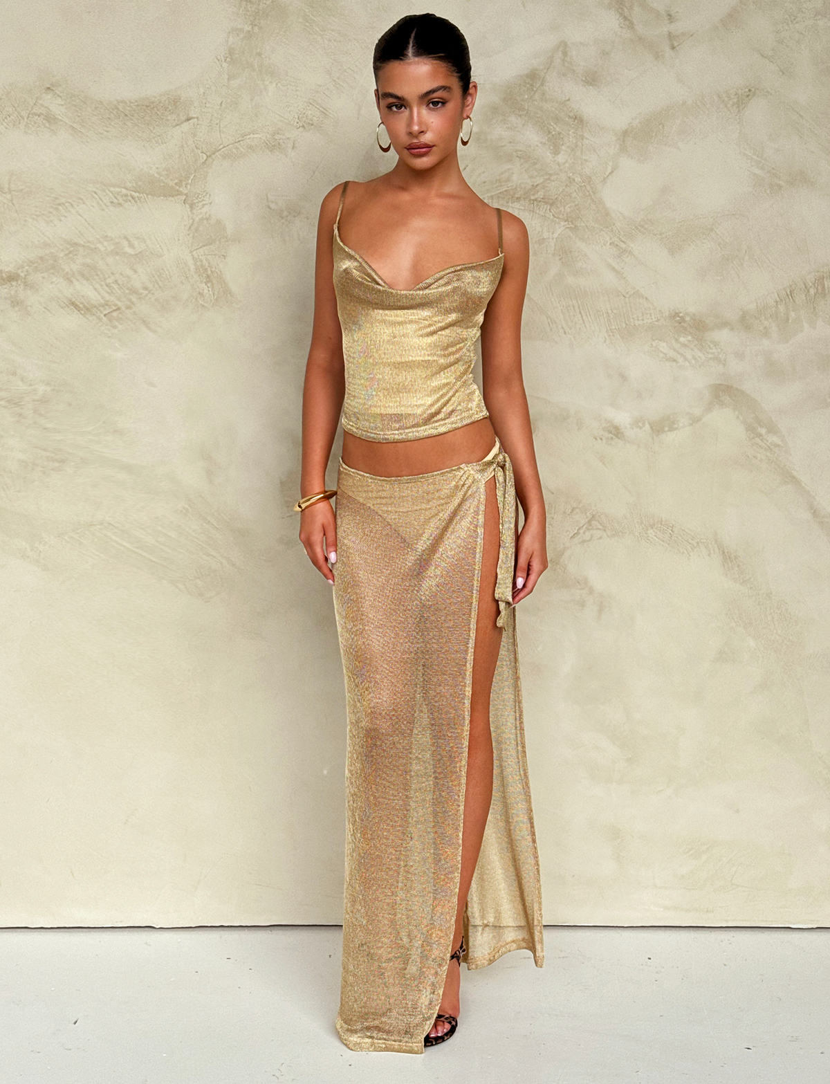 A woman poses against a textured beige wall in the TIGERMIST WYNNE SKIRT - GOLD, featuring a shimmery metallic finish and wrap detail. She pairs it with a draped top, hoop earrings, bracelets, and black heels for an effortlessly chic look.