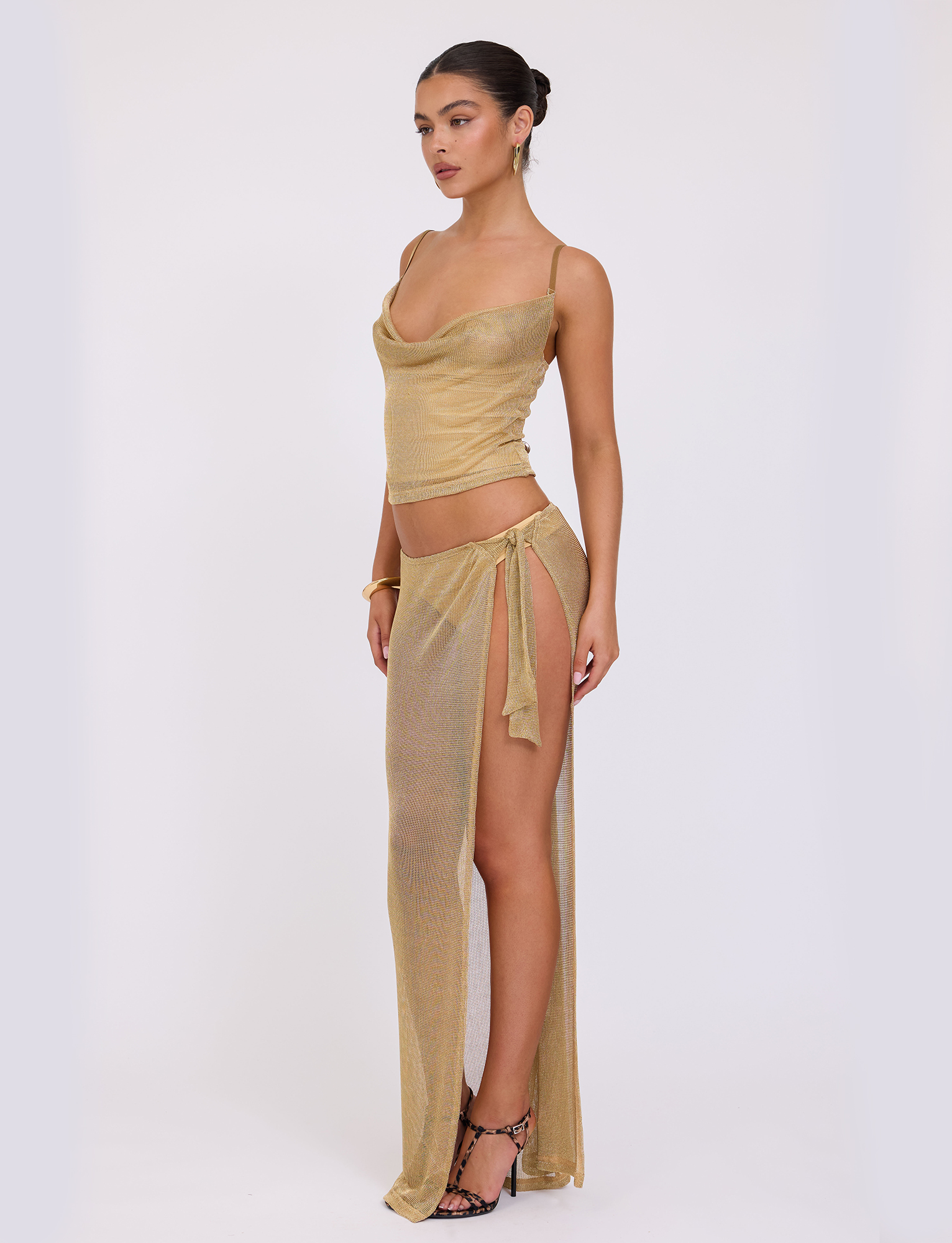 A woman poses against a white background wearing the TIGERMIST WYNNE SKIRT in gold, a metallic two-piece outfit with a cowl neck top and high side slit wrap skirt, styled with hoop earrings, a bracelet, and strappy heels.
