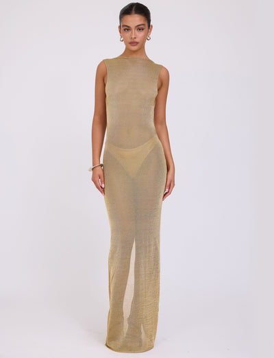 A woman stands against a plain white background, wearing the ZAELA DRESS - GOLD by TIGERMIST. The sheer, sleeveless, floor-length gown is form-fitting. She has her hair pulled back and accessorizes with round earrings and a bracelet.