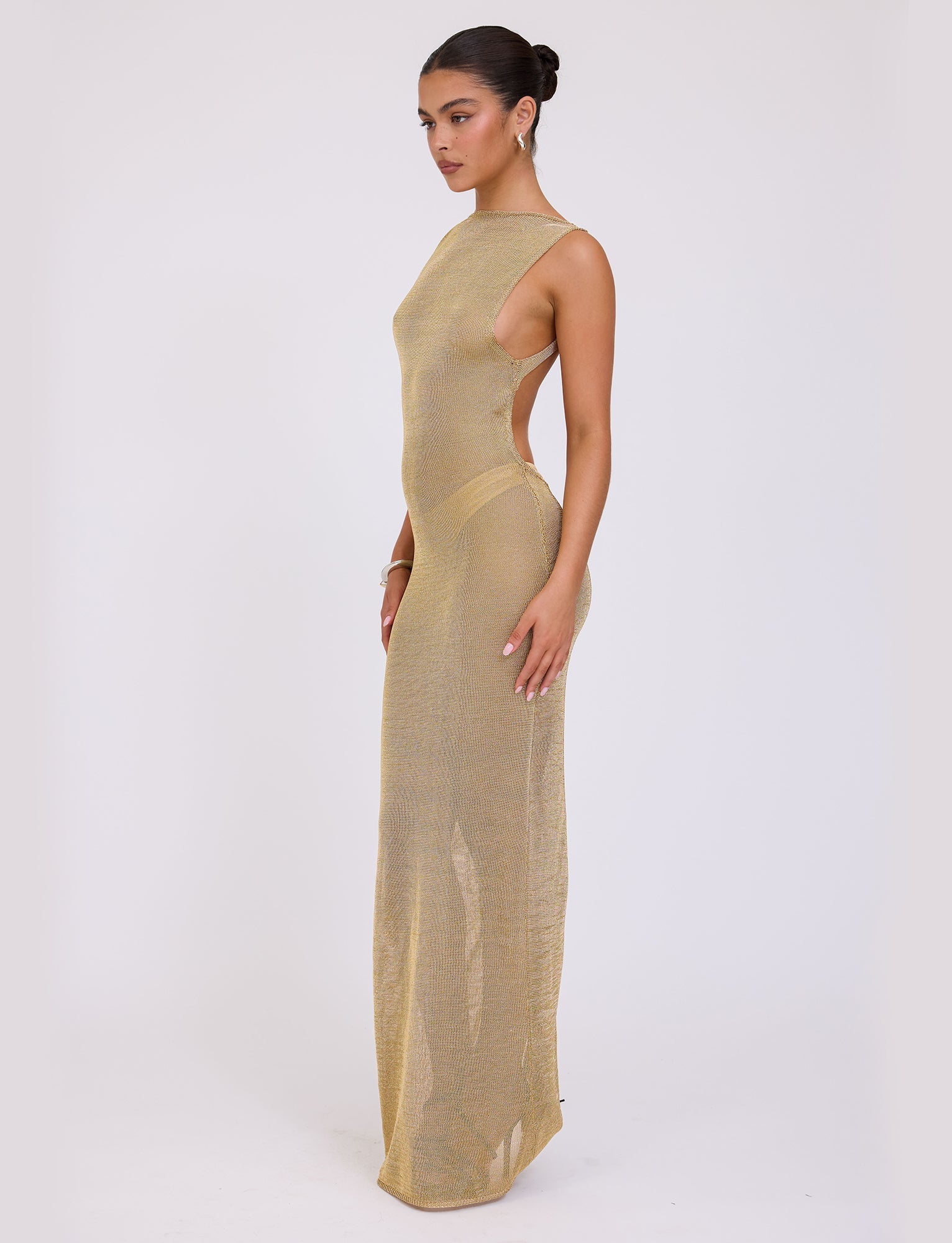 A woman stands against a plain white background, modeling the ZAELA DRESS - GOLD from TIGERMIST. The sheer, form-fitting dress features a high neckline, sleeveless design, and side cutouts. Her hair is styled in a sleek bun, and she gazes softly to the side while wearing hoop earrings and a bracelet.