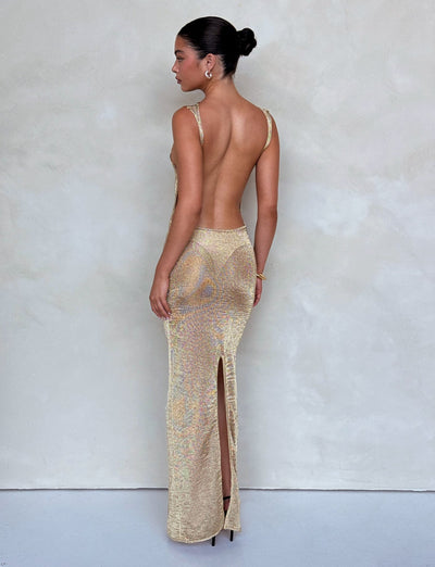 A woman with dark hair in a neat bun is wearing the ZAELA DRESS - GOLD from TIGERMIST. The glittering, backless gown features thin straps and a high slit at the back. She is standing and facing away from the camera, showcasing the dress's shimmering texture against a plain, light-colored background.