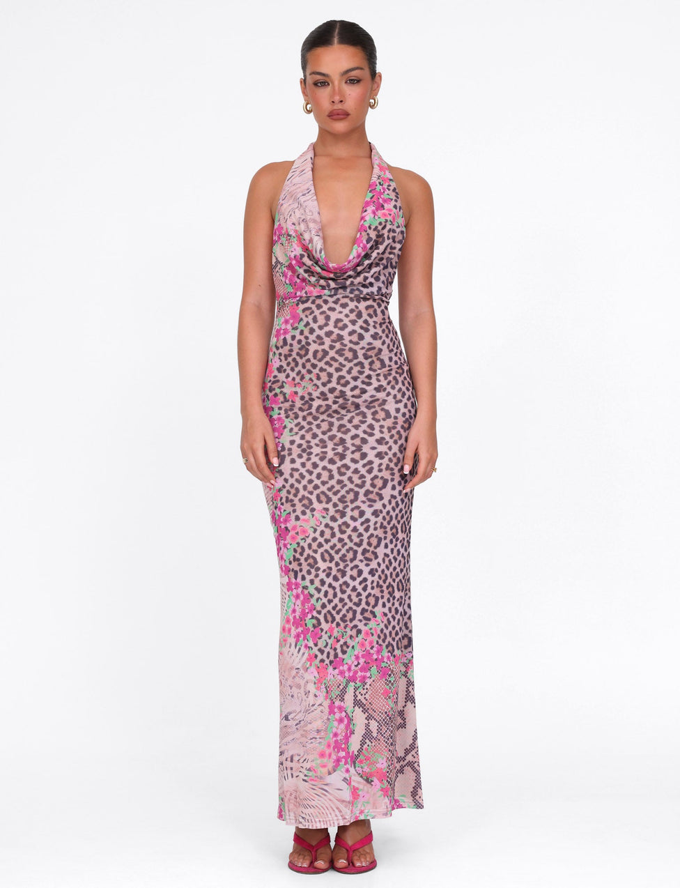 A woman faces forward in the TIGERMIST CATALINA MAXI DRESS in Animal Floral Border, her hair pulled back and styled with pink heels and subtle earrings, set against a plain white background.
