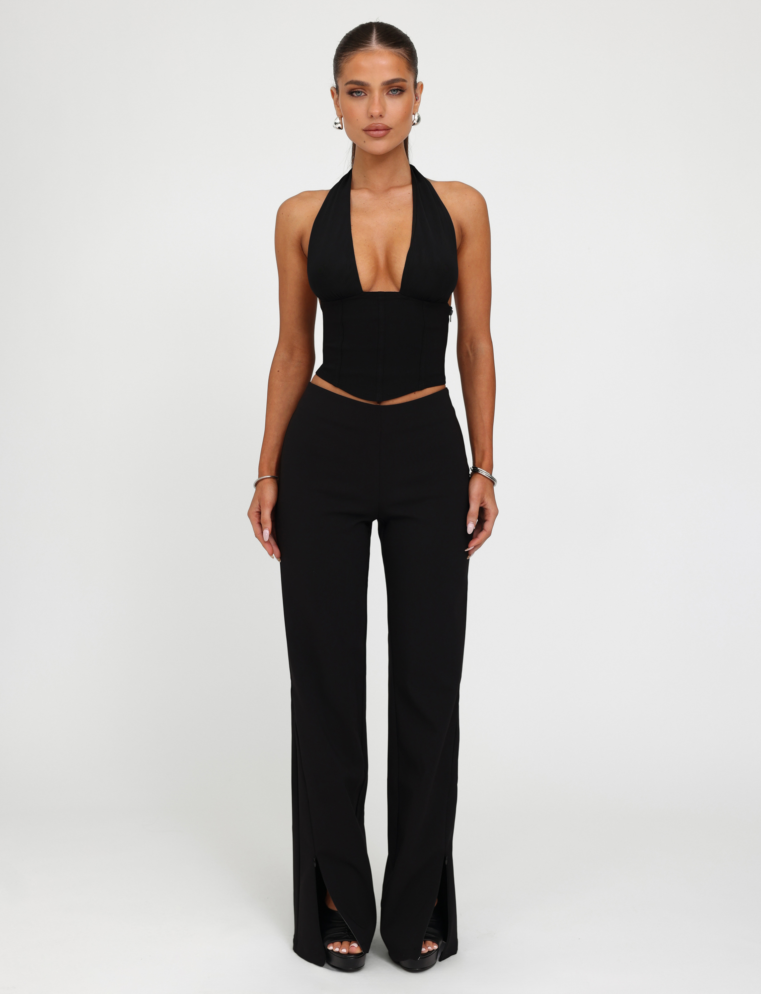 A woman faces forward against a white background, wearing the TIGERMIST JANECE TOP in black with high-waisted black pants featuring front slits, styled with earrings, bracelets, and rings.