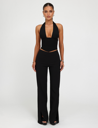 A woman faces forward against a white background, wearing the TIGERMIST JANECE TOP in black with high-waisted black pants featuring front slits, styled with earrings, bracelets, and rings.