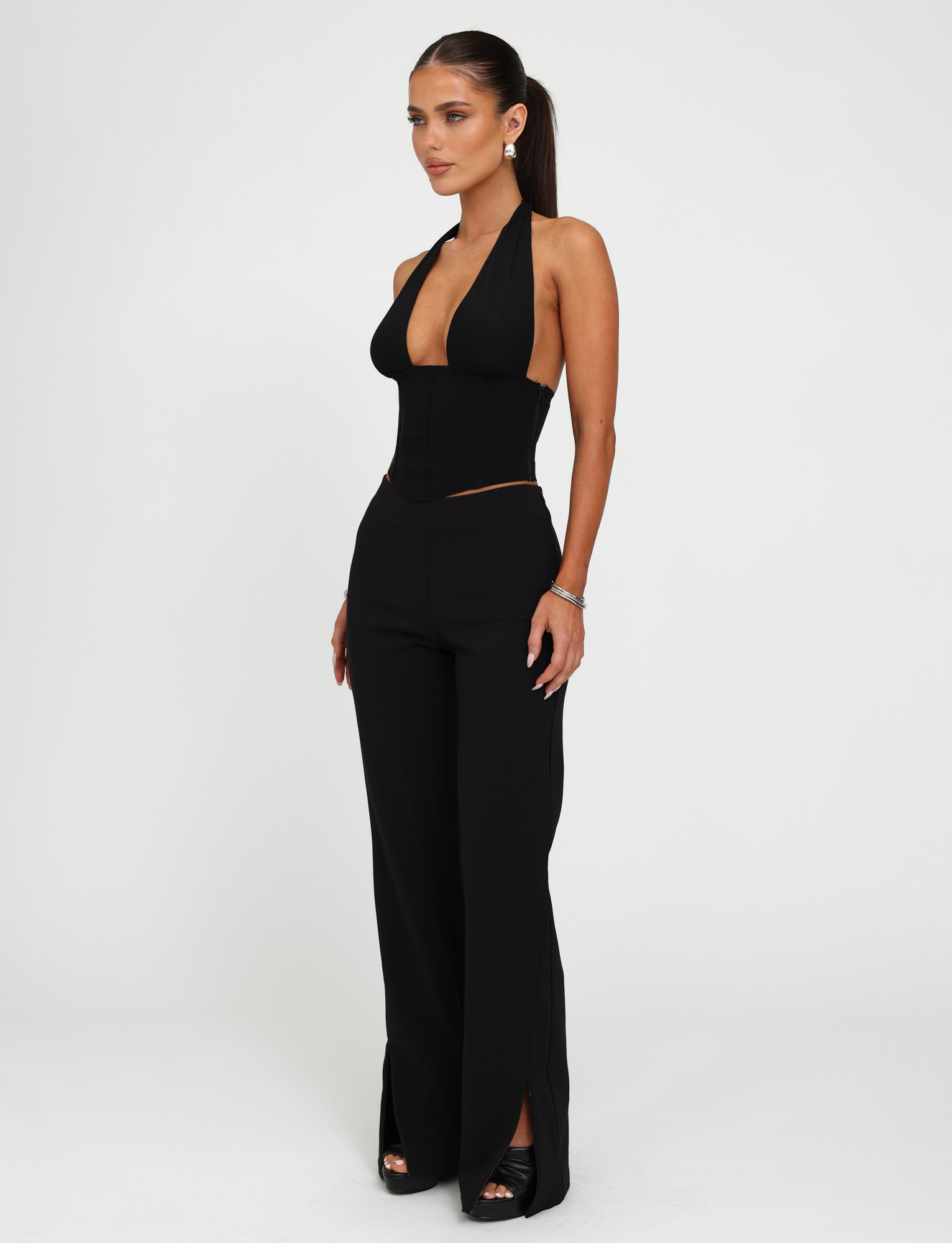 A woman wears the TIGERMIST JANECE TOP - BLACK, a sleeveless halter corset with a deep neckline, paired with high-waisted black pants featuring front hem slits. Her hair is in a ponytail and she completes the look with black open-toe heels.