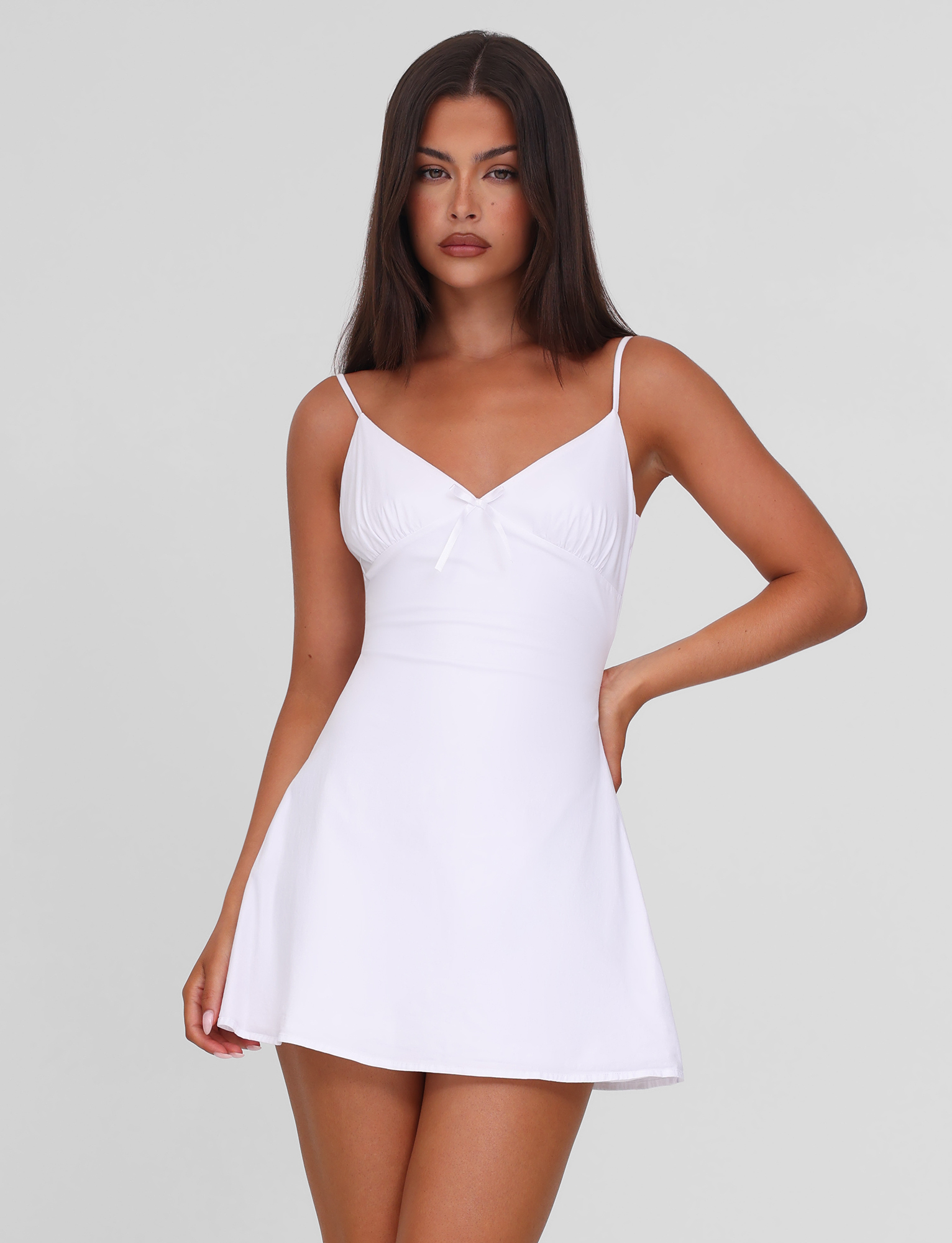 A woman with long dark hair models the TIGERMIST WINNIE MINI DRESS in white, a sleeveless, fitted style with adjustable straps and a flared hem, posing against a plain light gray background.