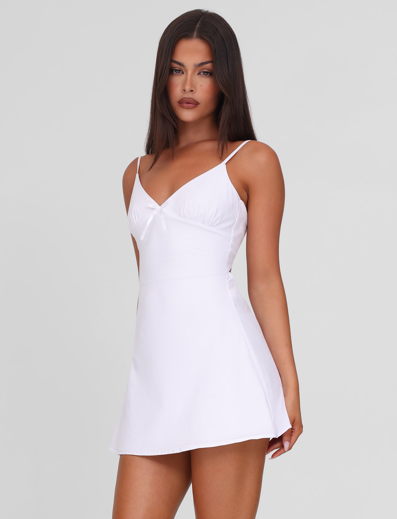 A woman with straight dark hair wears the TIGERMIST WINNIE MINI DRESS in white, featuring adjustable straps and a small neckline bow, standing against a plain light gray background.