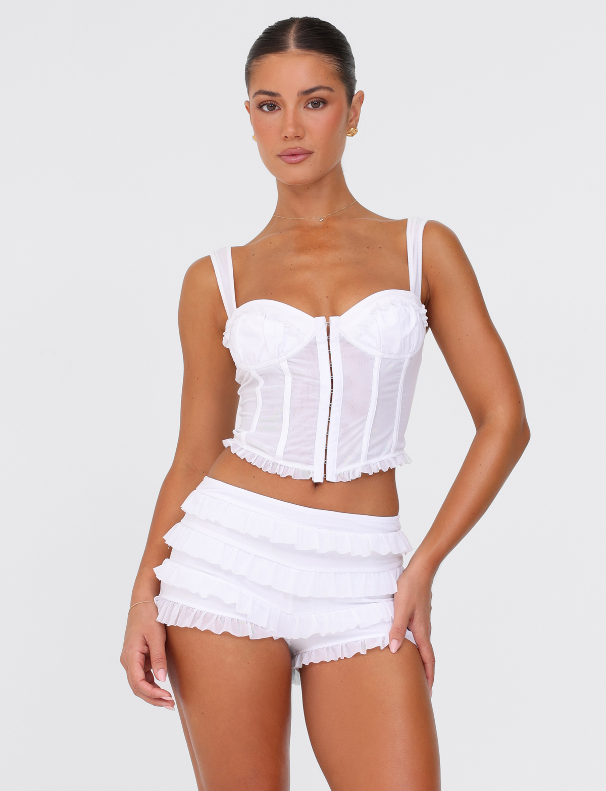 A woman poses confidently with her hair pulled back, wearing the TIGERMIST GINNY TOP in optic white—a corset-style top with a front zipper—paired with matching high-waisted ruffle shorts against a plain white background.