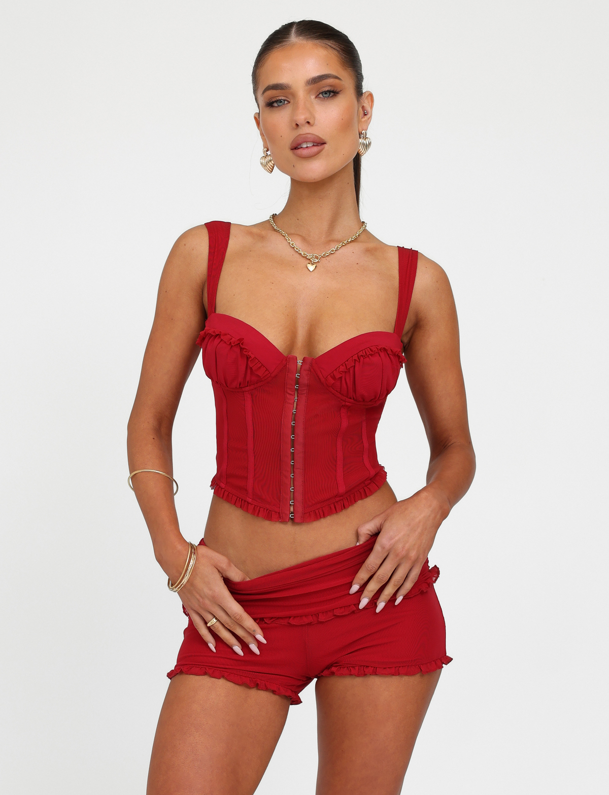 Model poses against a white backdrop wearing the TIGERMIST GINNY TOP - RED in cherry red, featuring a sweetheart neckline and ruffled details, styled with matching red shorts and gold jewelry for a chic look.