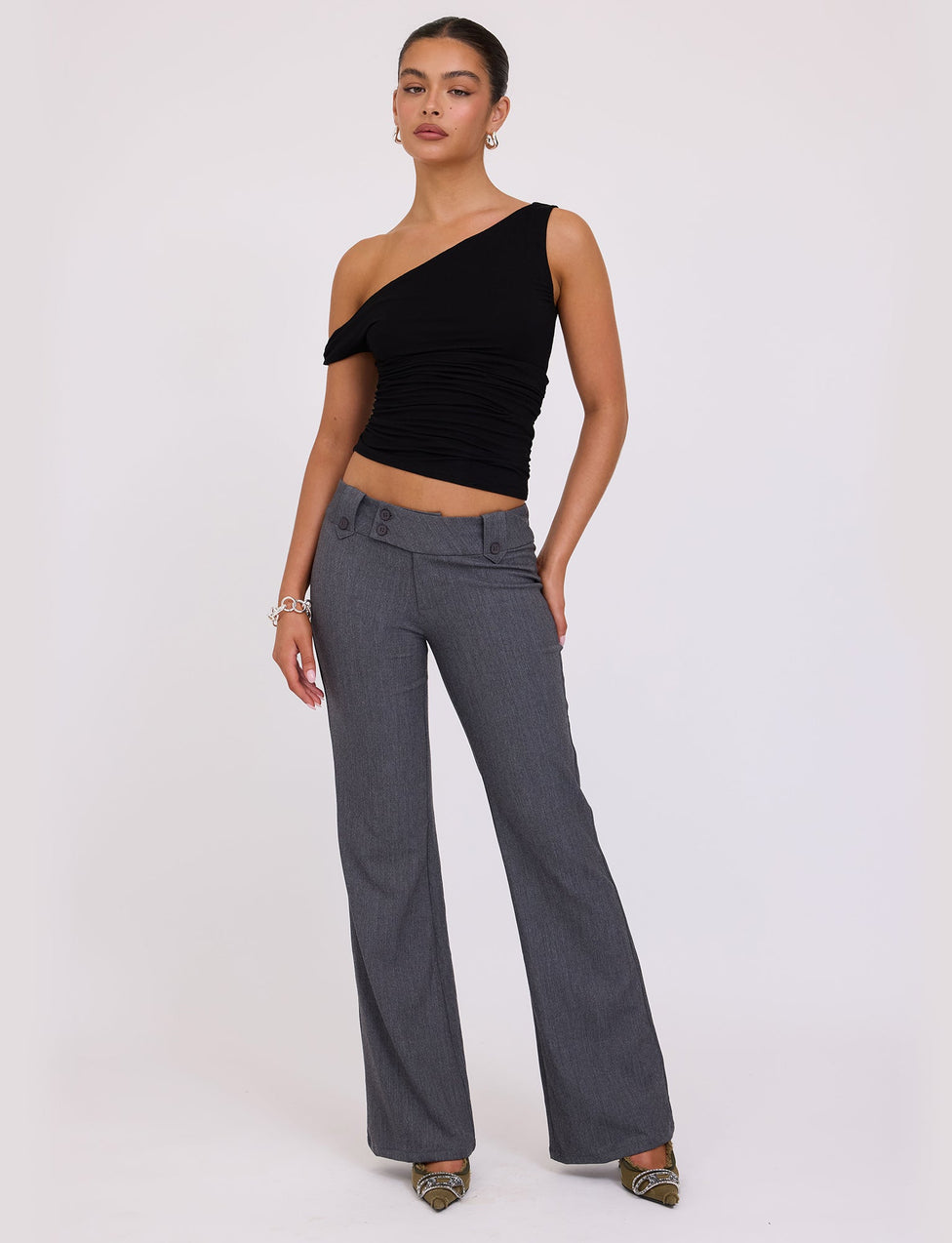 A woman is standing against a plain background, wearing the KADENCE TOP - BLACK from TIGERMIST and high-waisted gray trousers. She has her hair tied back and accessorizes with a bracelet and dangling earrings. She is wearing pointed-toe shoes and posing with one hand on her hip.