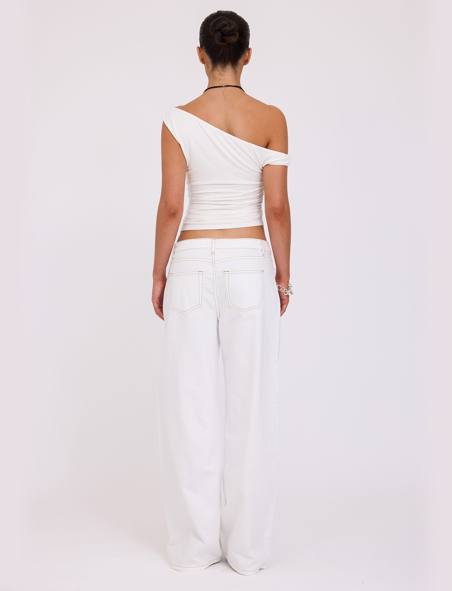 A person is standing with their back to the camera, stylishly dressed in the Tiger Mist FELIX JEAN - WHITE, featuring a one-shoulder top and high-waisted, wide-leg pants. Their hair is styled in a neat bun, complemented by a bracelet and necklace.