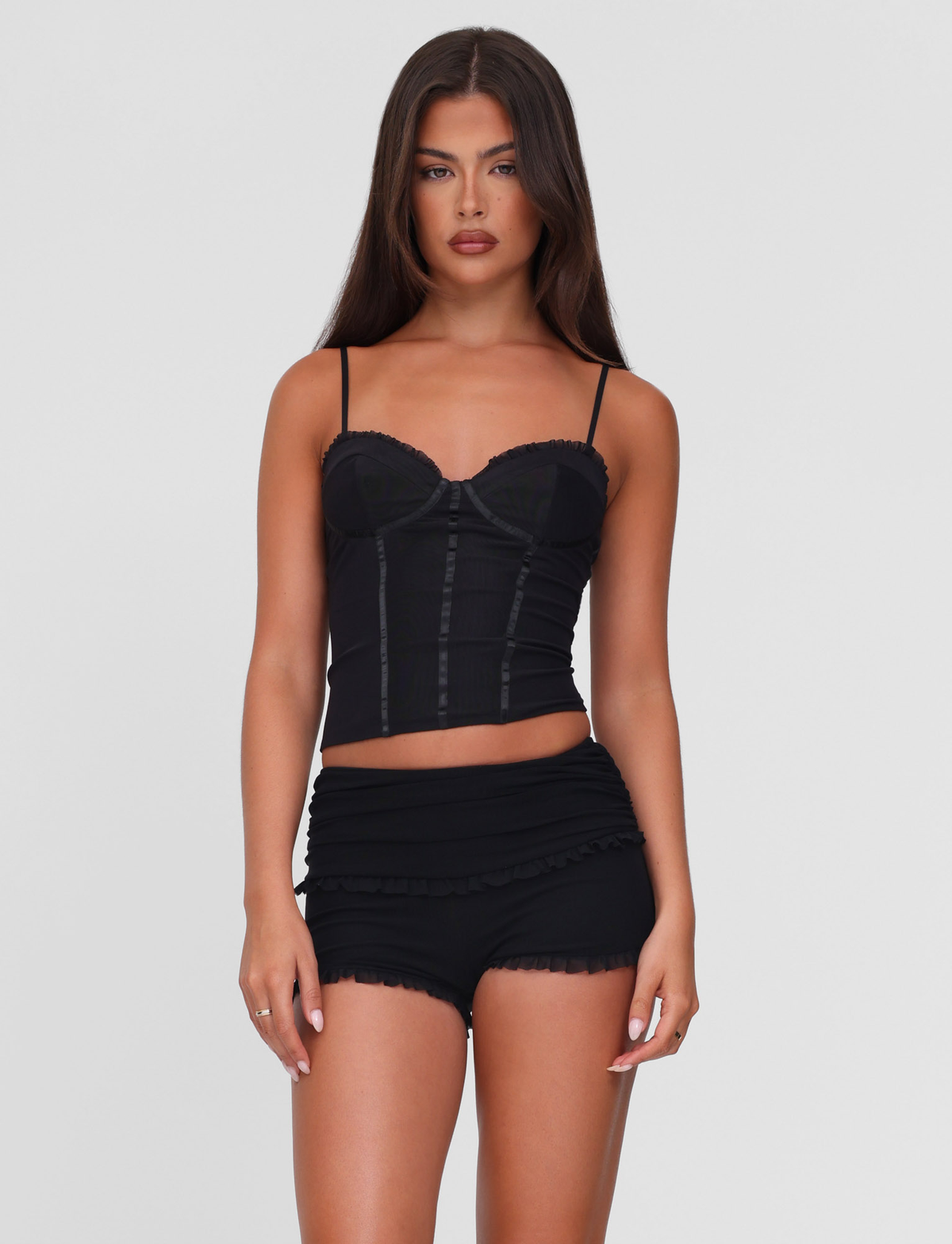 A woman with long brown hair wears the TIGERMIST GINNY SHORT - BLACK and a fitted black camisole, standing against a plain light gray background.