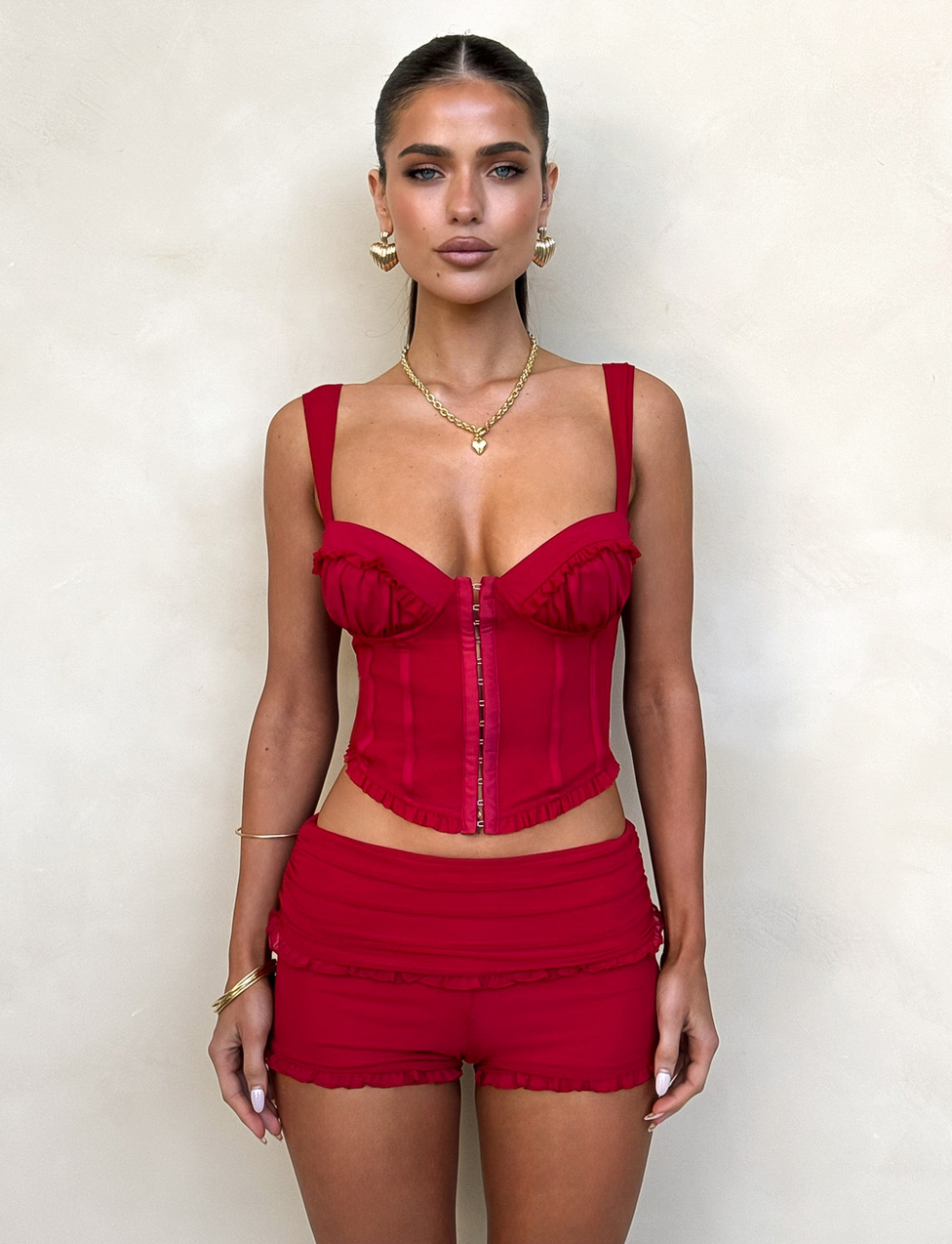 A woman poses against a beige wall in the TIGERMIST GINNY SHORT - RED mini dress, featuring ruffle details. She wears her hair pulled back with gold jewelry and exudes a neutral, confident vibe.