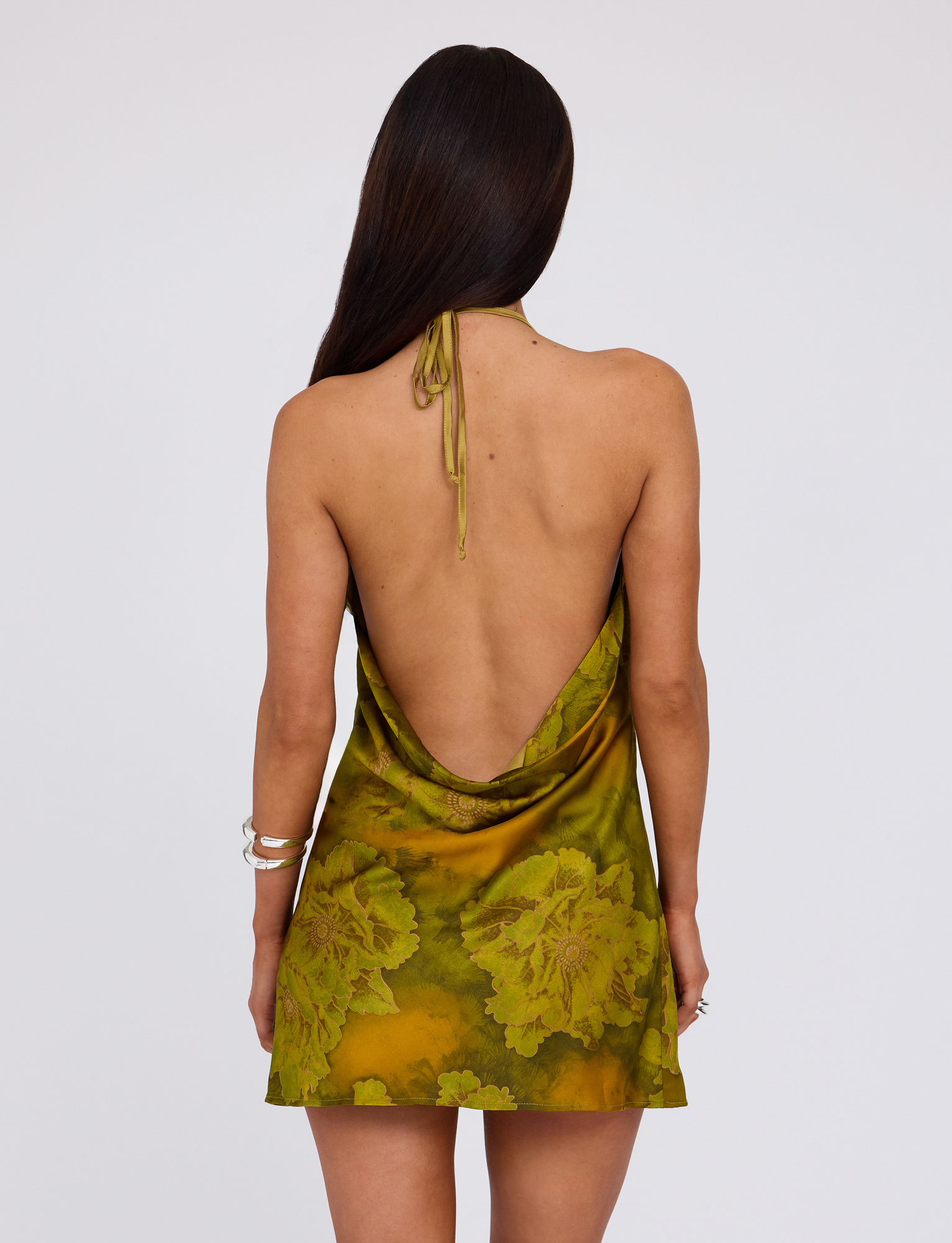 A woman with long dark hair, seen from behind, wears the TIGERMIST MONROE DRESS - GREEN: WATER COLOUR FLORAL, a backless halter style. She accessorizes with silver bracelets and stands against a plain light background.