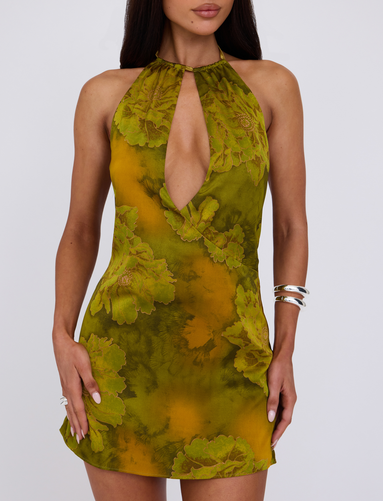 A woman wears the TIGERMIST MONROE DRESS - GREEN : WATER COLOUR FLORAL, a sleeveless mini dress featuring a deep plunging neckline and keyhole detail, accessorized with silver bangles and rings against a plain background.