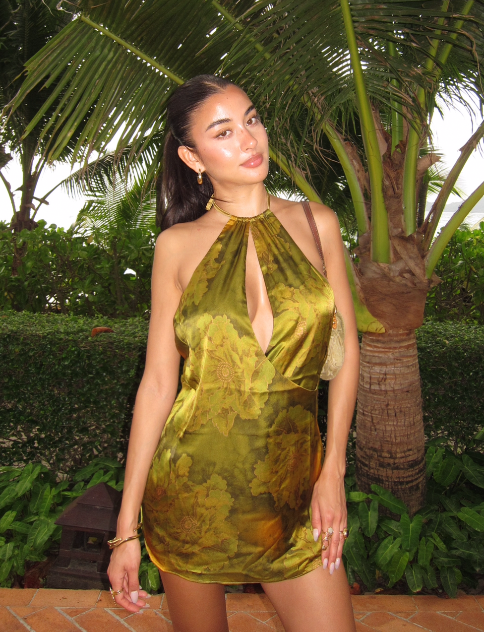 A woman poses outdoors among tropical plants in the TIGERMIST MONROE DRESS - GREEN : WATER COLOUR FLORAL, featuring a shiny olive-green floral print and halter neckline. Her long dark hair is pulled back and she wears gold jewelry.