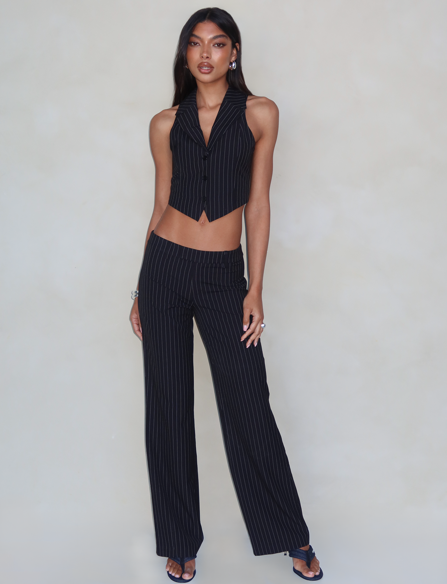 A person with long dark hair stands confidently against a neutral background, wearing the TIGERMIST TAMARA PANT - BLACK : PINSTRIPE and a matching pinstriped, sleeveless suit vest. The wide-legged pants feature a straight-leg silhouette and a mid-rise waist. Paired with open-toed black heels, their relaxed pose includes one hand in their pocket and the other by their side.