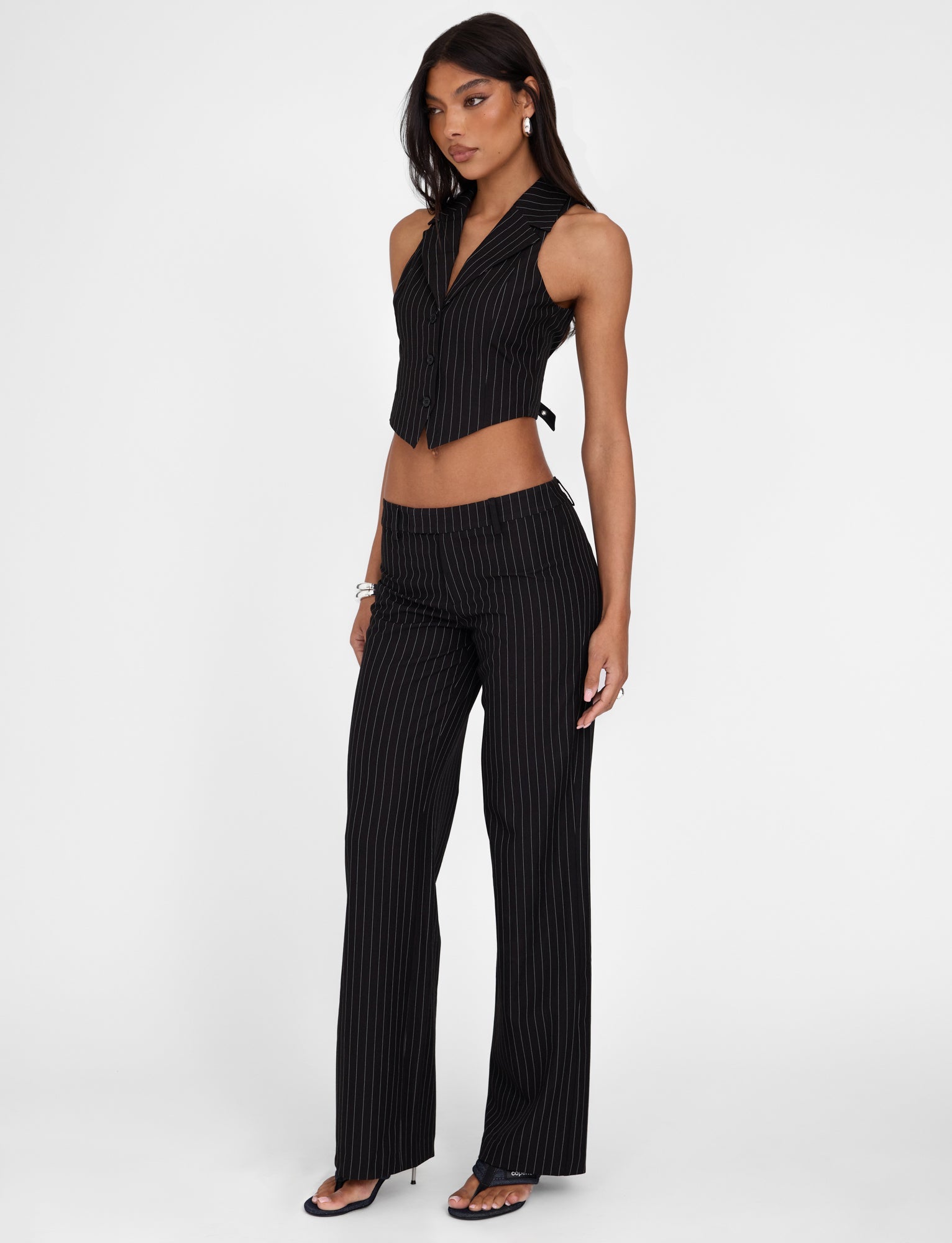 A woman stands confidently in the TIGERMIST TAMARA PANT - BLACK : PINSTRIPE two-piece outfit: a sleeveless vest and mid-rise waist, straight leg silhouette trousers. She has long dark hair, hoop earrings, bracelets on one wrist, and is wearing open-toe heels. The background is plain white.