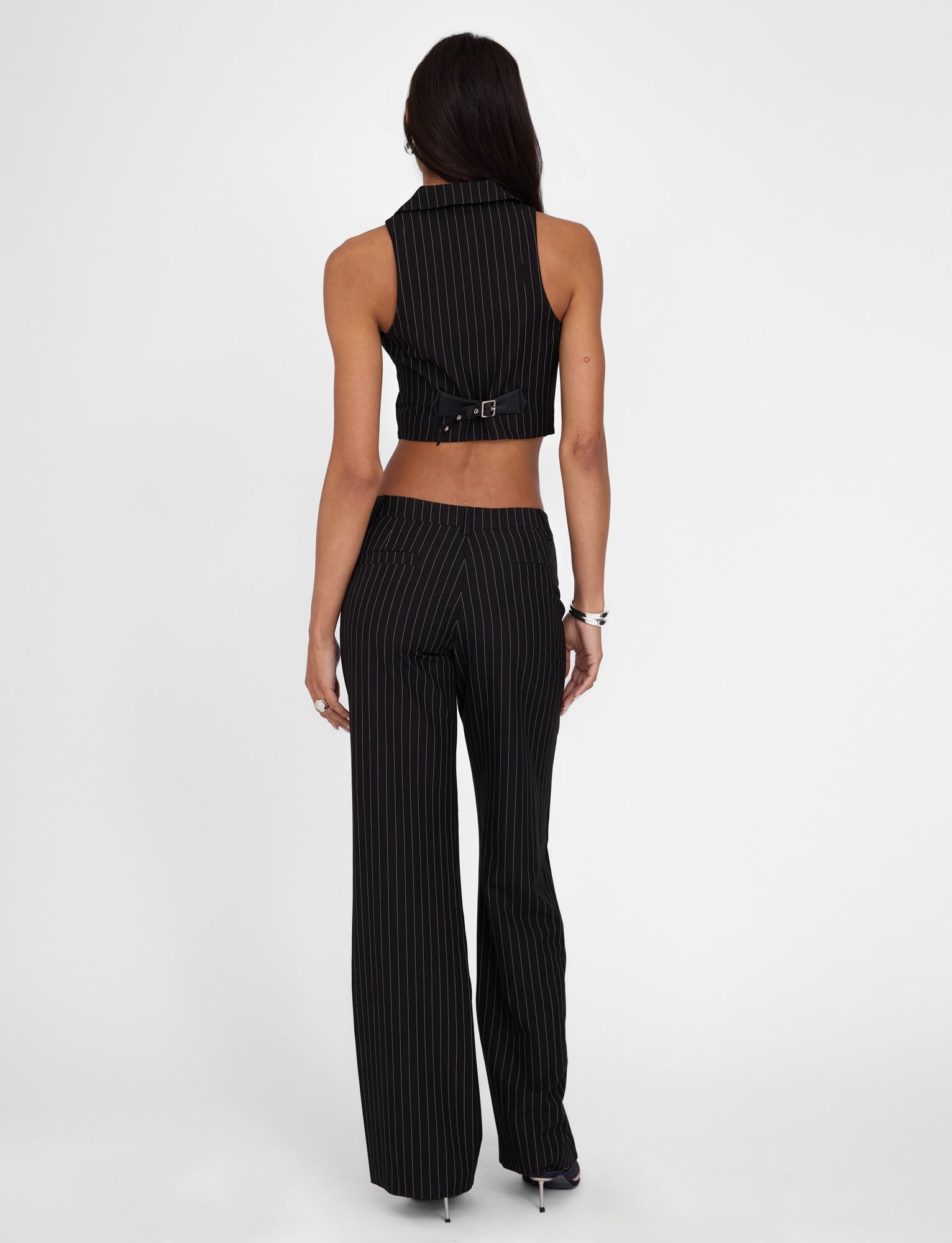 A woman stands with her back to the camera, showcasing the TAMARA PANT - BLACK : PINSTRIPE by TIGERMIST, paired with a matching black pinstripe crop top featuring a buckle at the back. She has long dark hair and is posed against a plain white background.