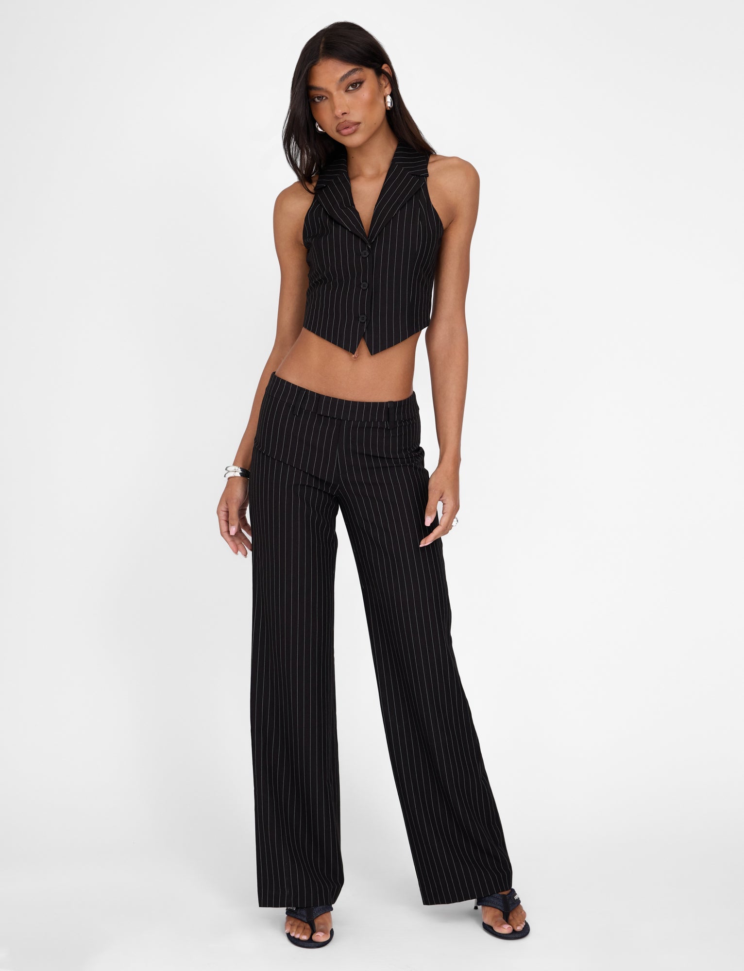 A woman strikes a confident pose in the TIGERMIST TAMARA PANT - BLACK : PINSTRIPE set, which includes a sleeveless crop vest and high-waisted, straight-leg silhouette trousers. She complements her look with bracelets, earrings, and heeled sandals while standing against a plain white background.
