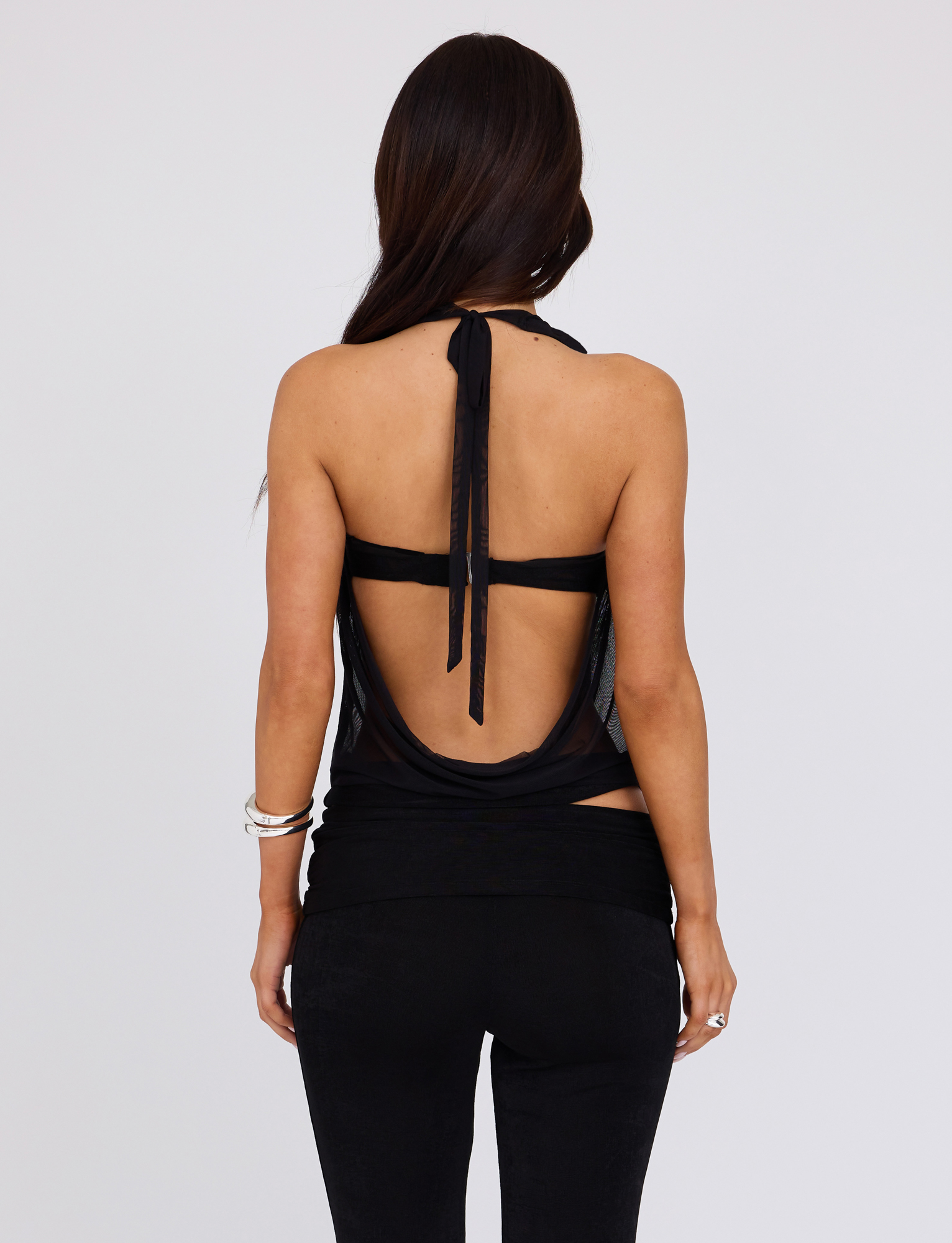 A person with long dark hair is shown from the back wearing the TIGERMIST MAKENA TWO-PIECE TOP in black and black pants, accessorized with bracelets and rings against a plain, light-colored background.