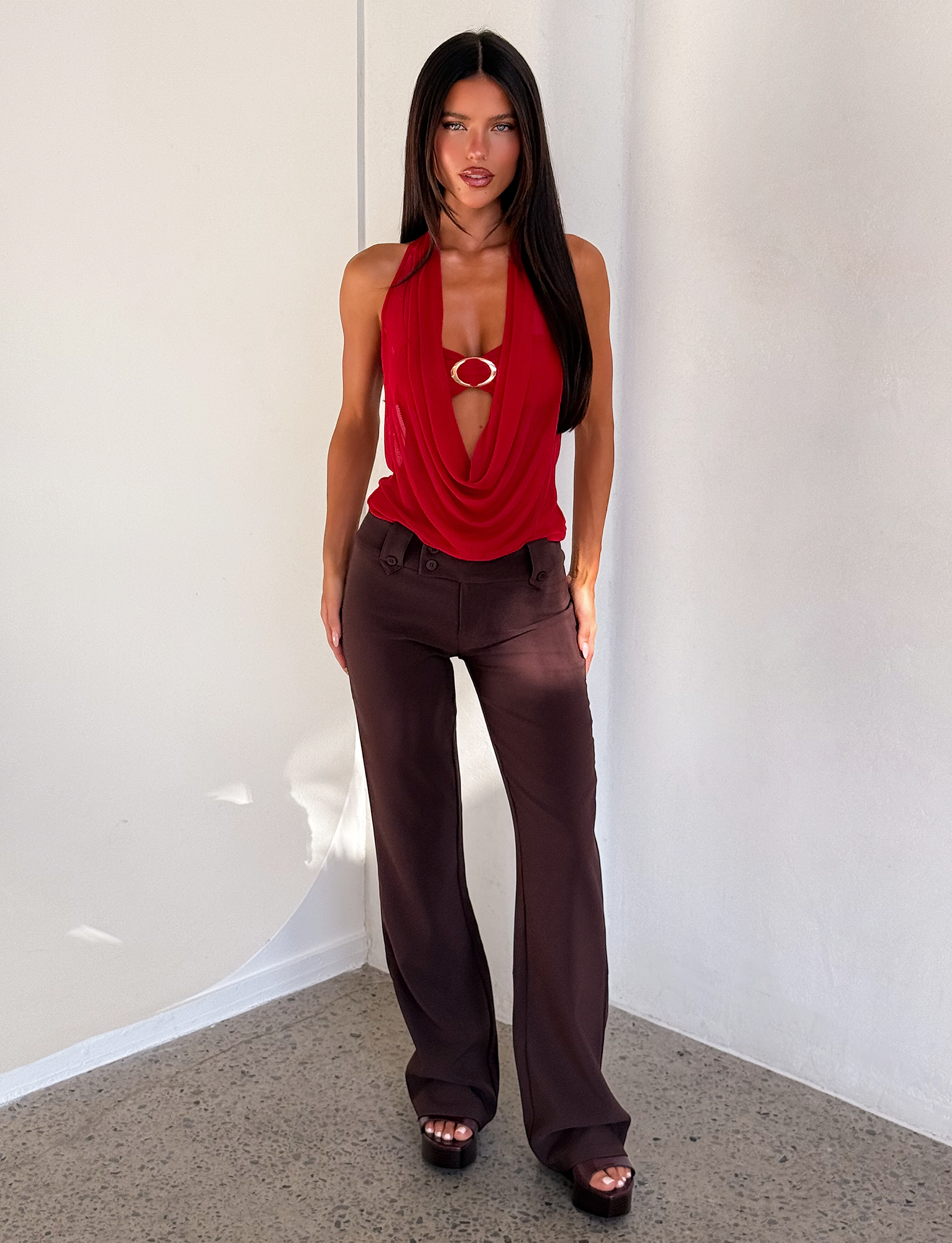 A woman with long dark hair stands indoors, facing forward against a white wall, wearing the TIGERMIST MAKENA TWO-PIECE TOP – RED, styled with high-waisted brown pants and platform sandals as light falls on her.