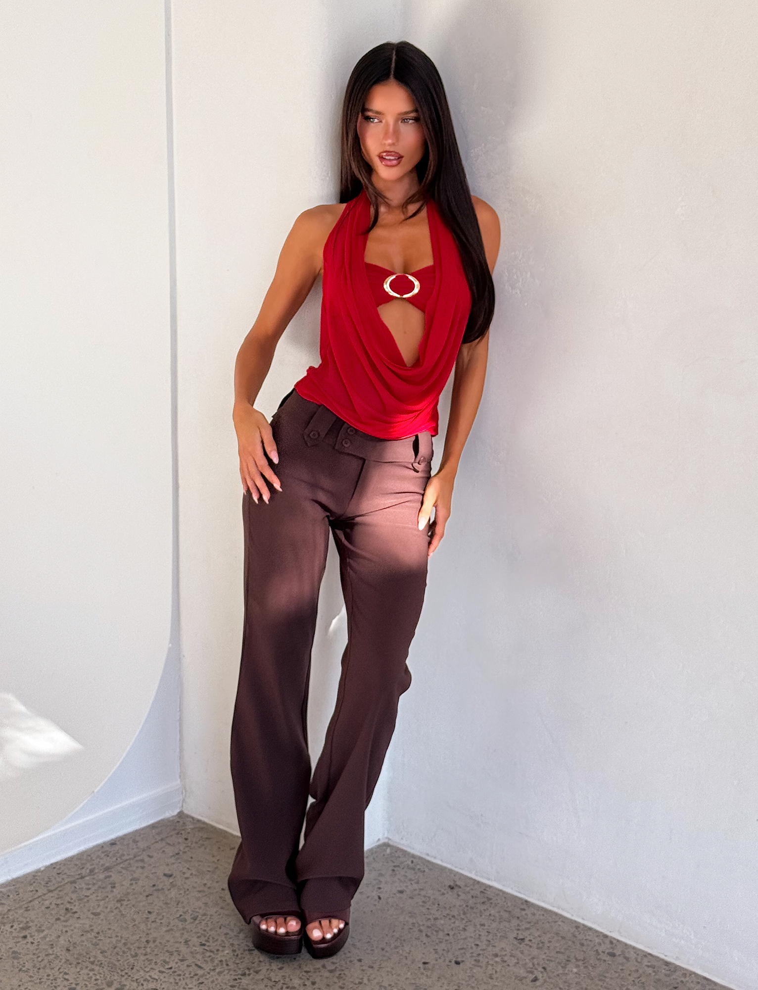 A woman with long dark hair wears the TIGERMIST MAKENA TWO-PIECE TOP - RED, featuring a circular metal detail, styled with brown high-waisted pants and platform sandals, standing against a light-colored wall on a concrete floor.
