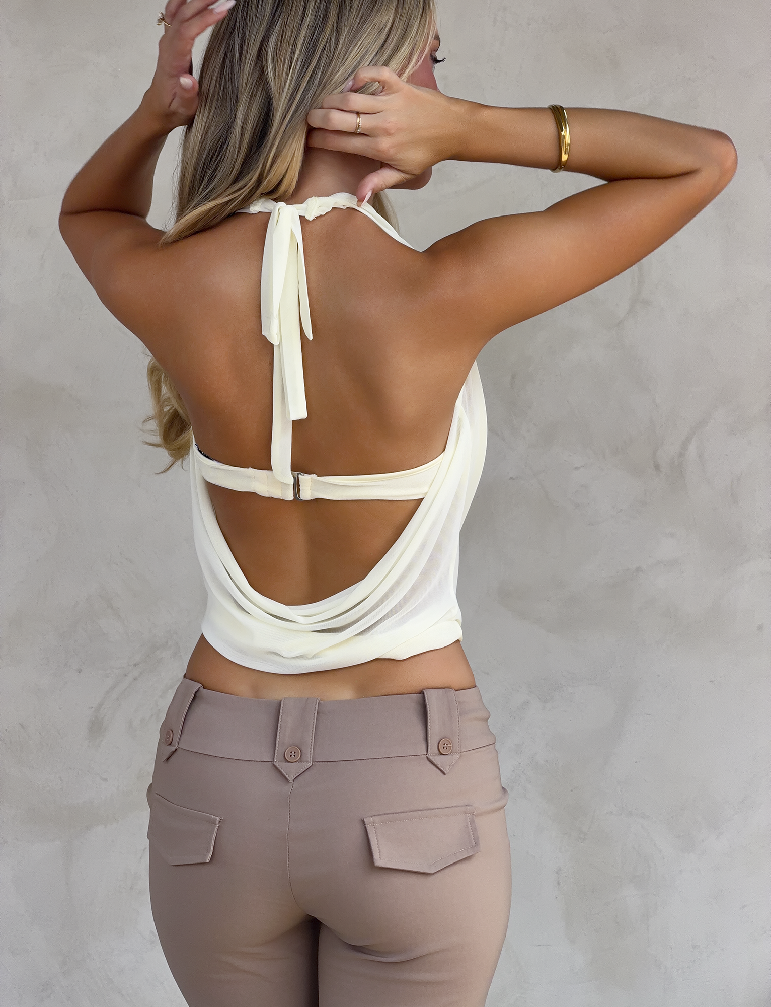 A woman with long blonde hair faces away, showing the draped open back and neck tie of the TIGERMIST MAKENA TWO-PIECE TOP in ivory. She pairs it with light brown pants and a gold bracelet, standing against a neutral wall.