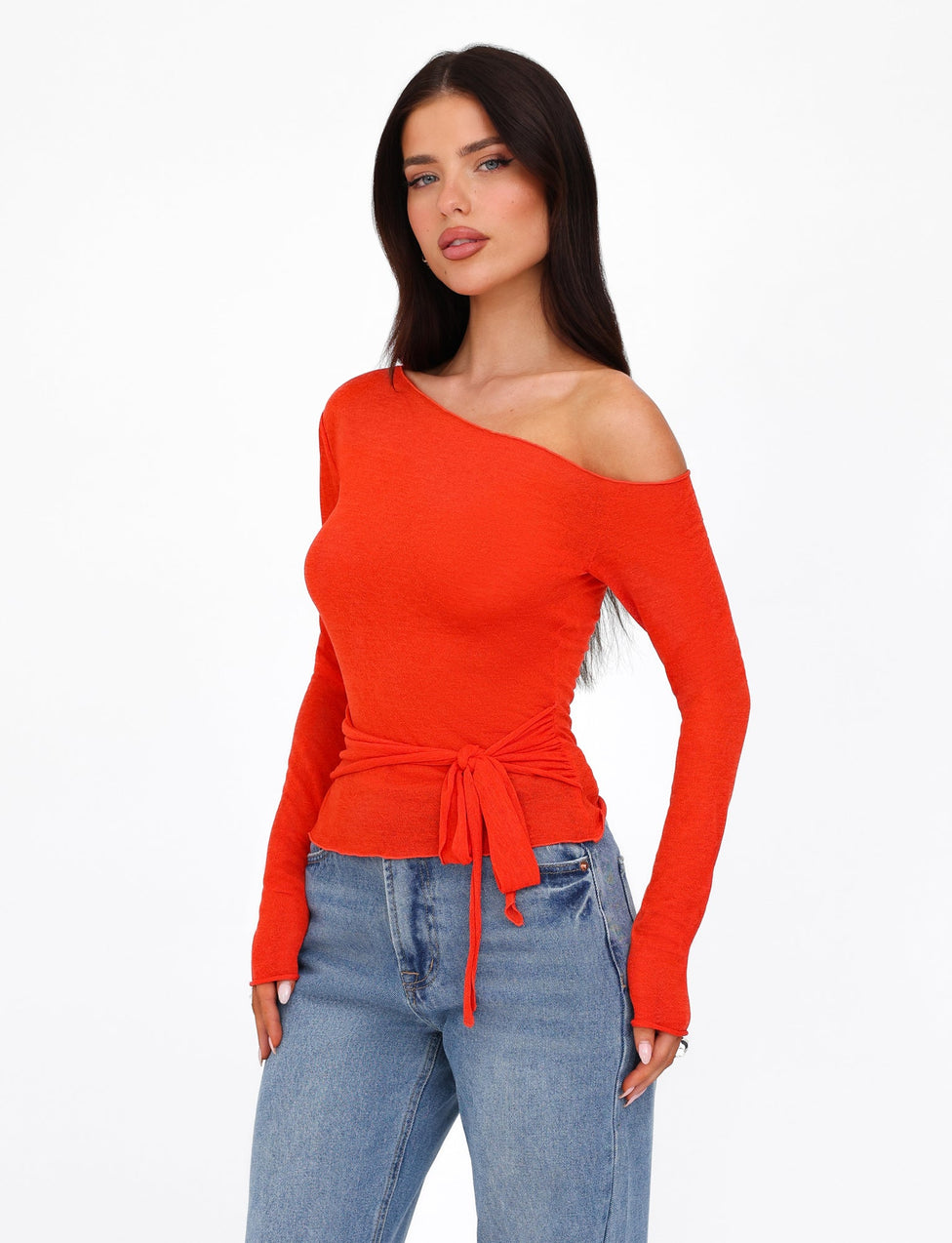 A woman with long dark hair is wearing the BRYNA JUMPER by TIGERMIST, a bright red off-shoulder, long-sleeve top featuring a tie waist, along with light blue jeans. The knit fabrication of the jumper complements her look as she gazes towards the camera with a neutral expression against a plain white background.