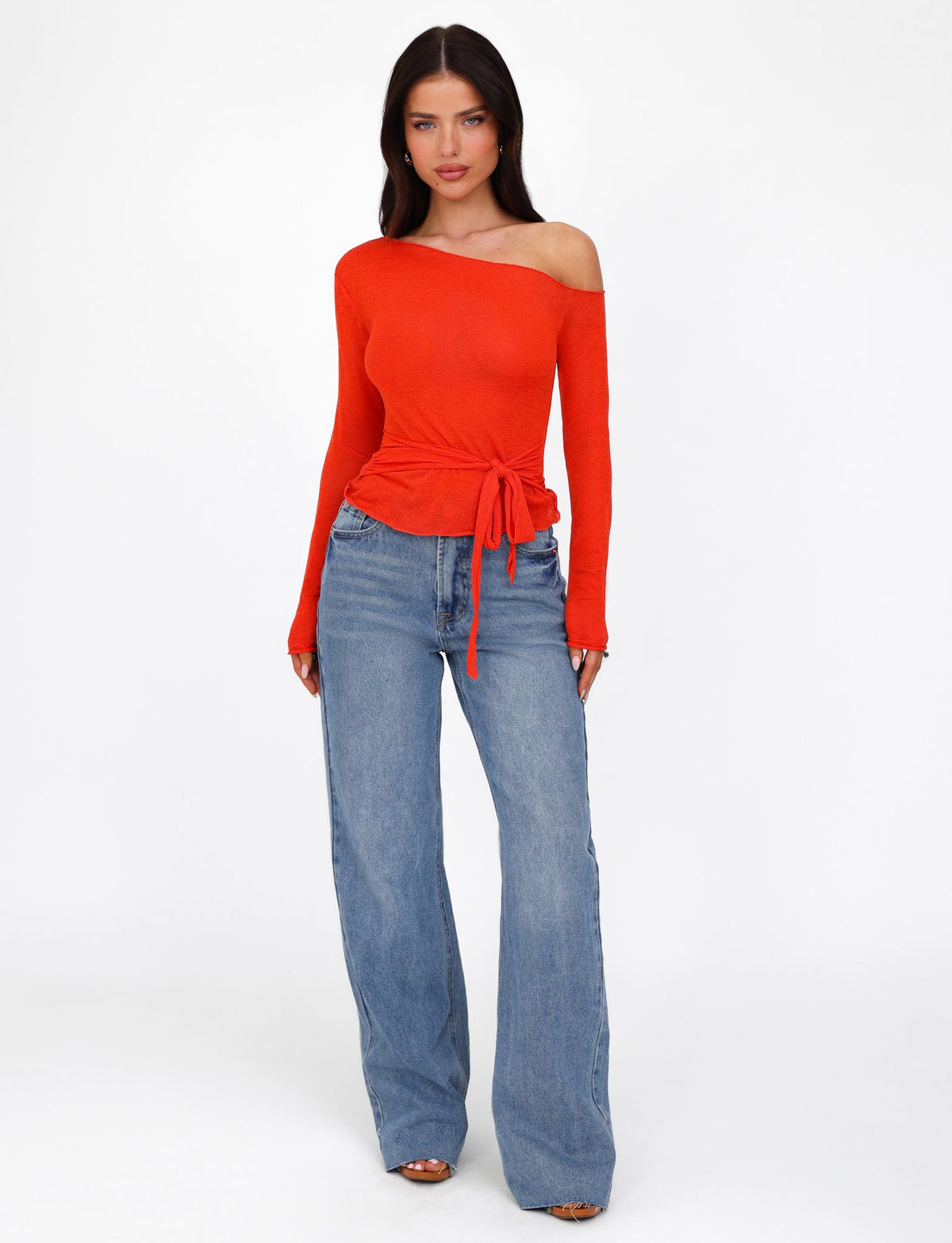 A person with long dark hair is wearing the TIGERMIST BRYNA JUMPER in red, which features an off-the-shoulder design and long sleeves, paired with loose-fitting blue jeans. They are standing against a plain white background, looking directly ahead with a neutral expression.