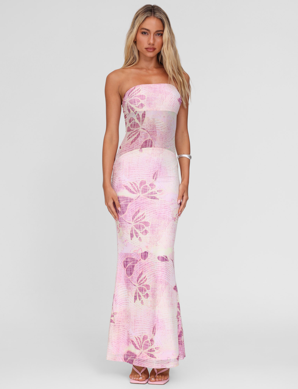 A woman with long, wavy blonde hair wears the TIGERMIST MORGAN MESH DRESS - GRUNGE FLORAL MAGENTA, a floor-length, strapless, fitted dress. She pairs it with sandals and a bracelet, posing against a plain light background.