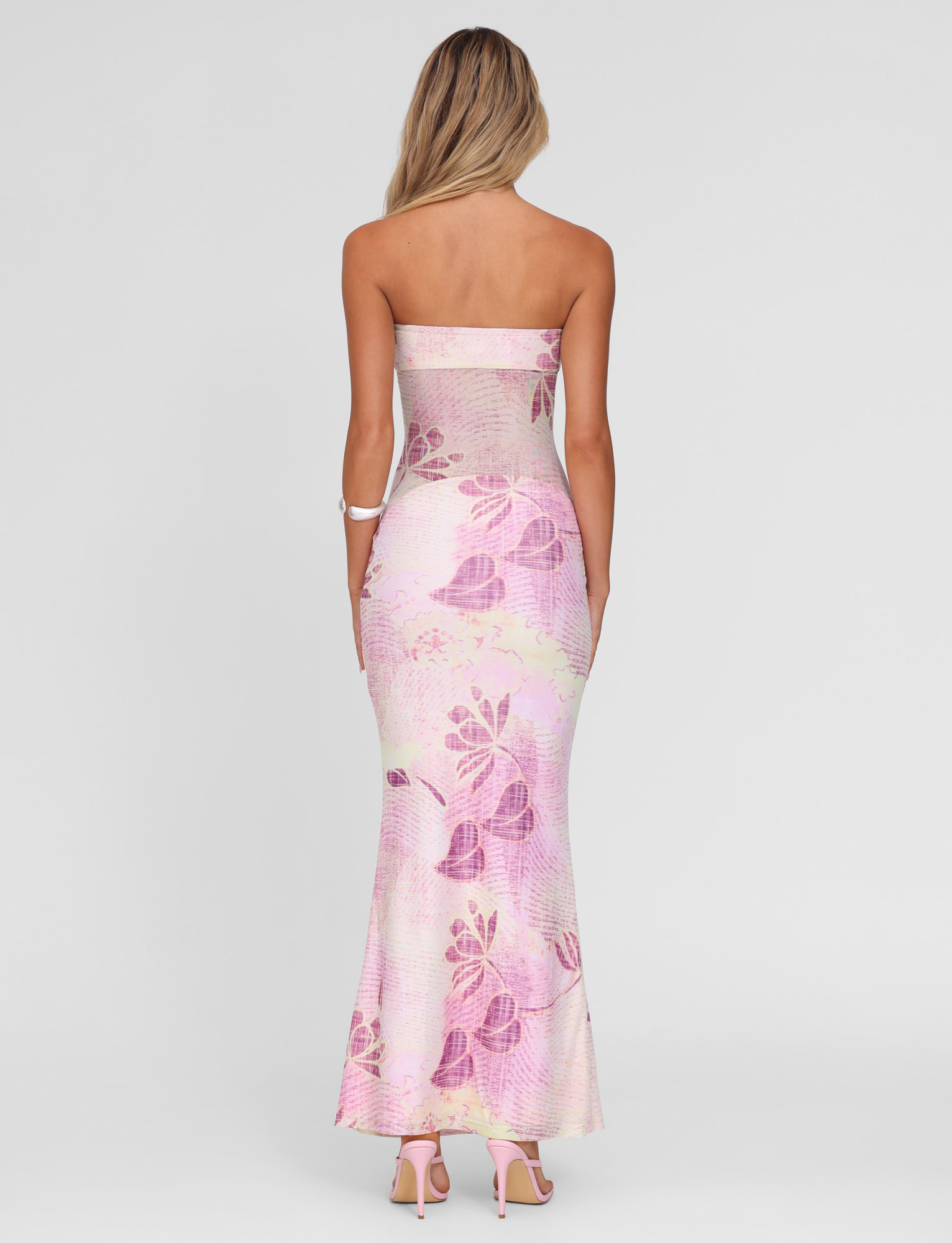 A woman with long blonde hair wears the TIGERMIST MORGAN MESH DRESS in Grunge Floral Magenta, featuring a strapless neckline. She faces away on a plain white background, styled with pink heels and a white bracelet.