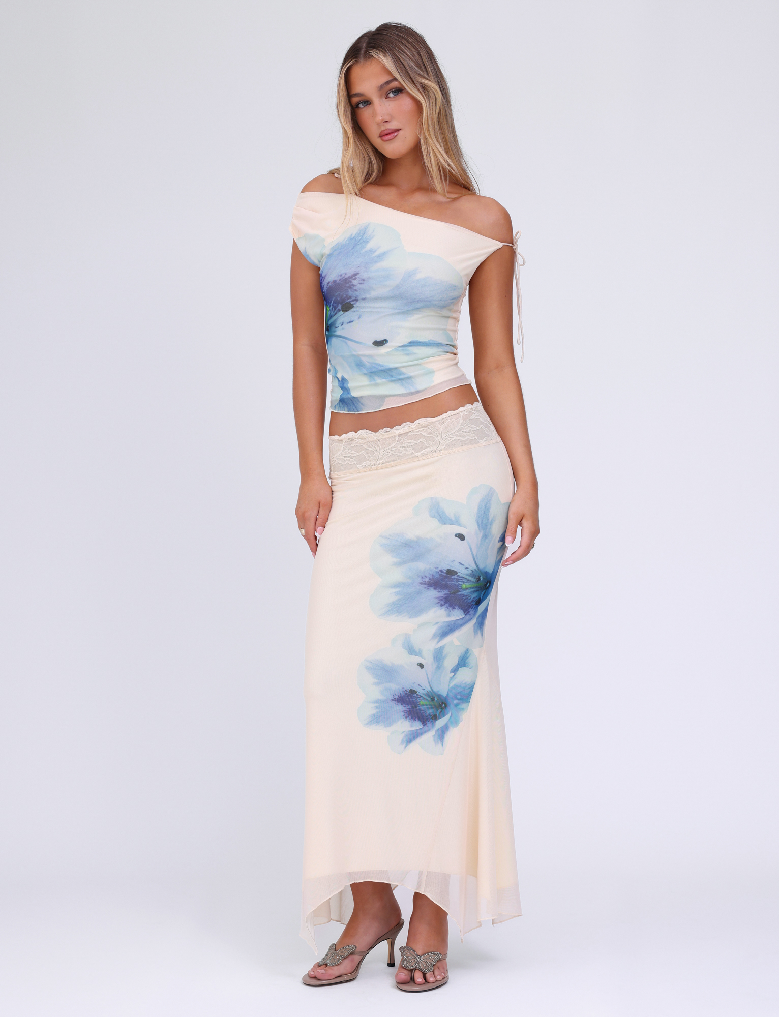 A woman with long blonde hair poses against a white background, wearing the TIGERMIST LILLIAN TOP in cream with a blue floral print and lace trim, paired with a matching skirt and gray sandals.