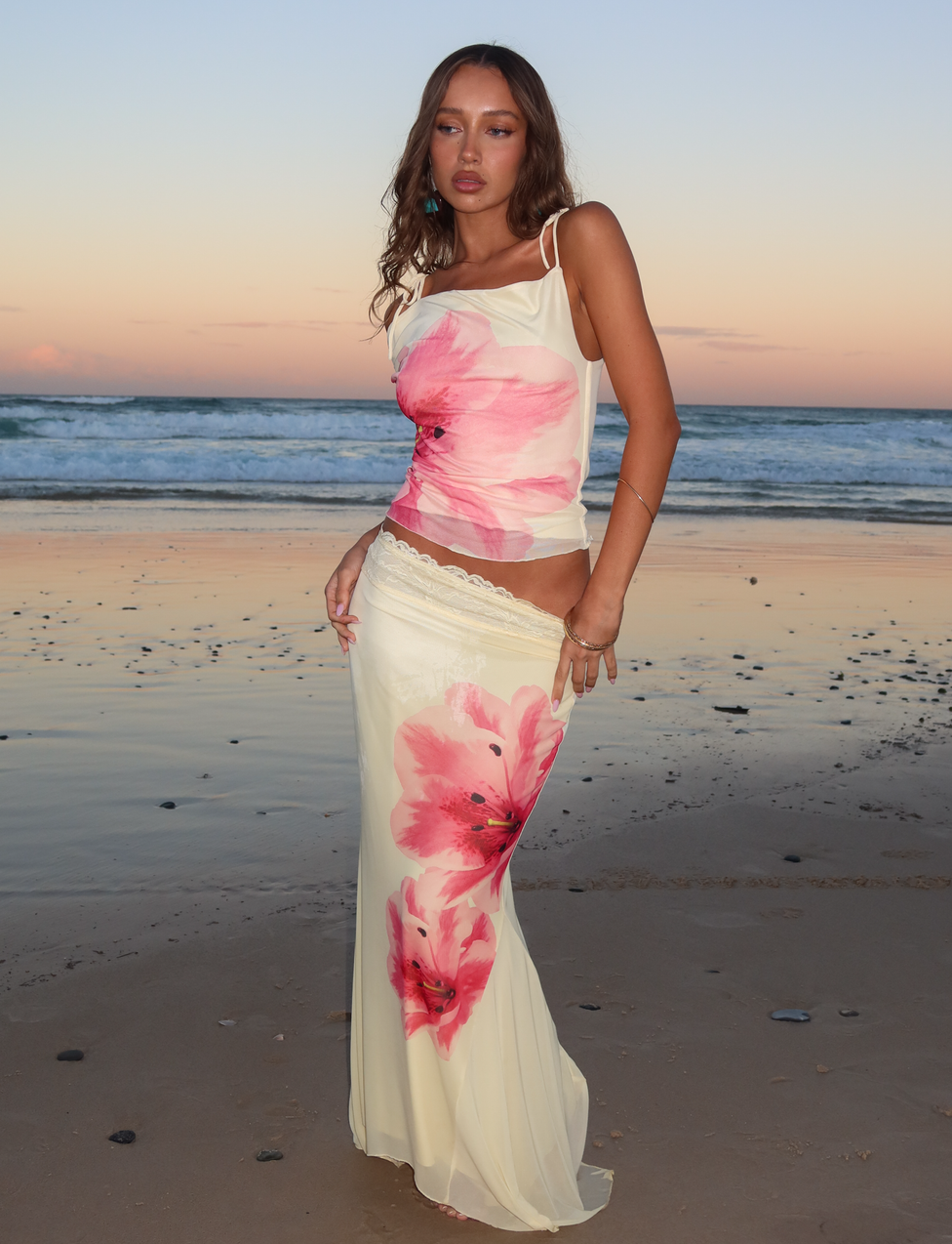 A woman stands on a beach at sunset wearing the CL - LILLIAN TOP by Tiger Mist USA, a cream-colored top with large pink floral prints, paired with a matching skirt. Ocean waves and a pastel sky create a serene backdrop.