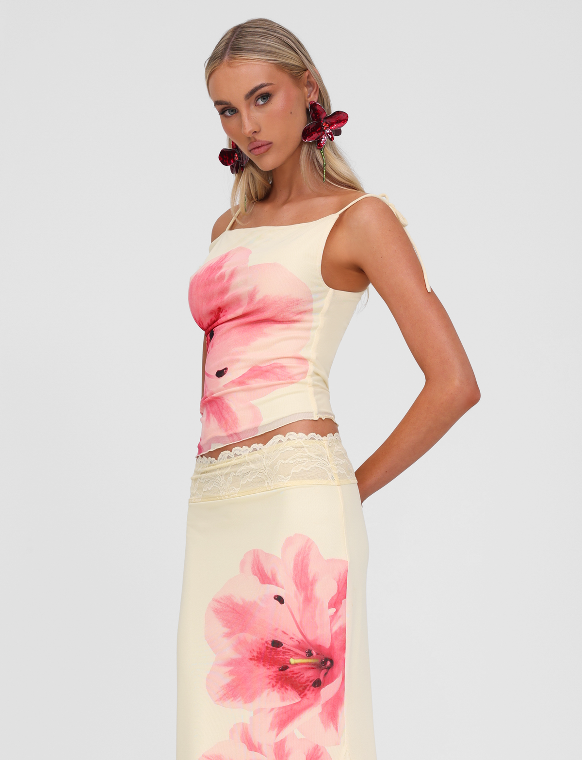 A woman with long blonde hair wears the TIGERMIST LILLIAN TOP in yellow, a strappy off-shoulder style, paired with a matching skirt. She accessorizes with bold red floral earrings and poses against a light gray background.