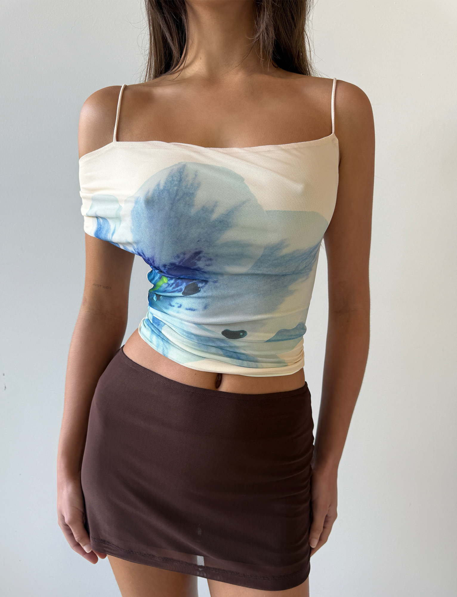 A woman wears the TIGERMIST LILLIAN TOP in white, cream, and floral, a sleeveless mesh top with blue flowers and thin straps, paired with a fitted high-waisted brown mini skirt, standing against a plain white background.