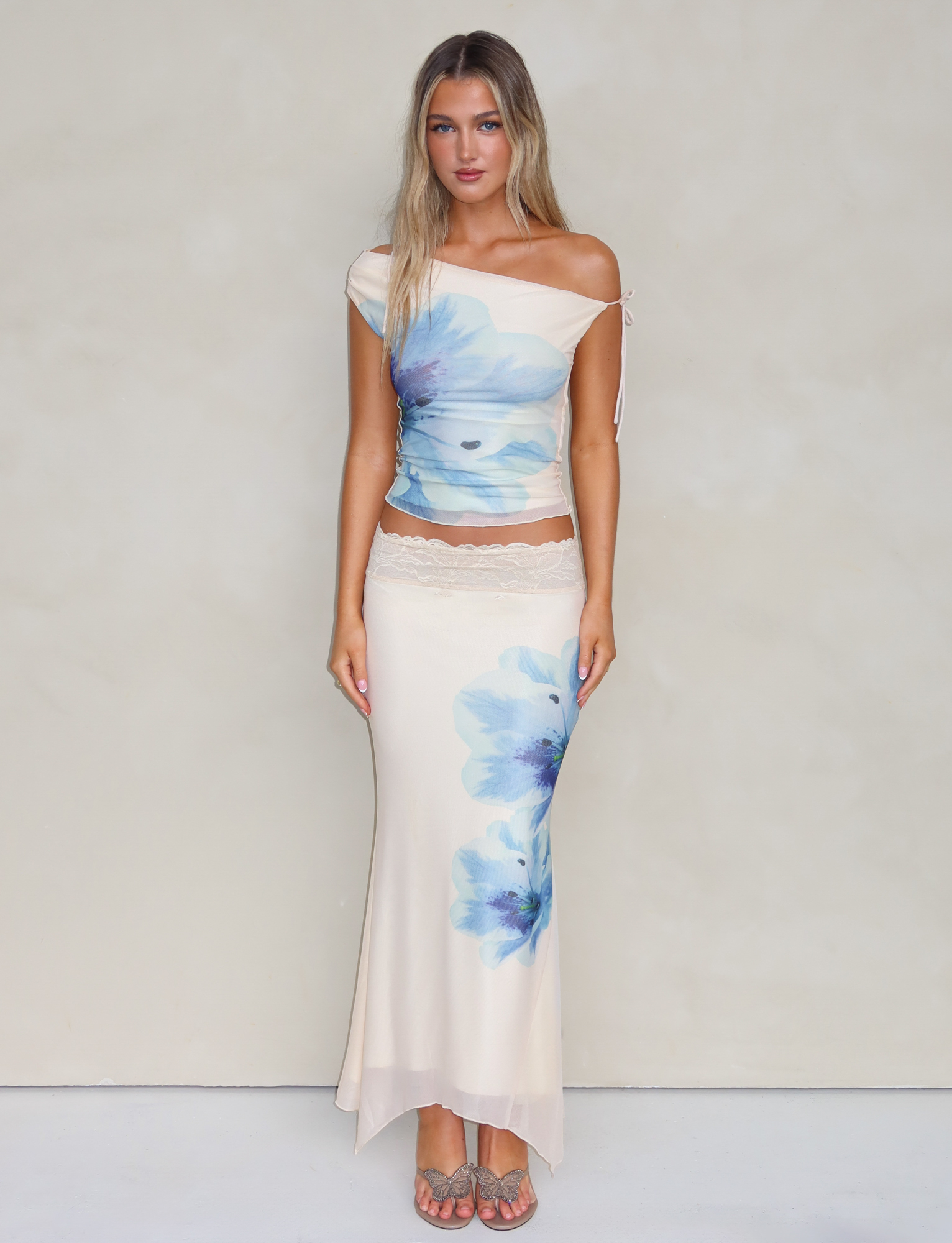 A woman stands against a plain light background wearing the TIGERMIST LILLIAN SKIRT in white/cream with blue floral prints. Her long blonde hair and neutral expression complete the look, while mesh fabric adds subtle texture.