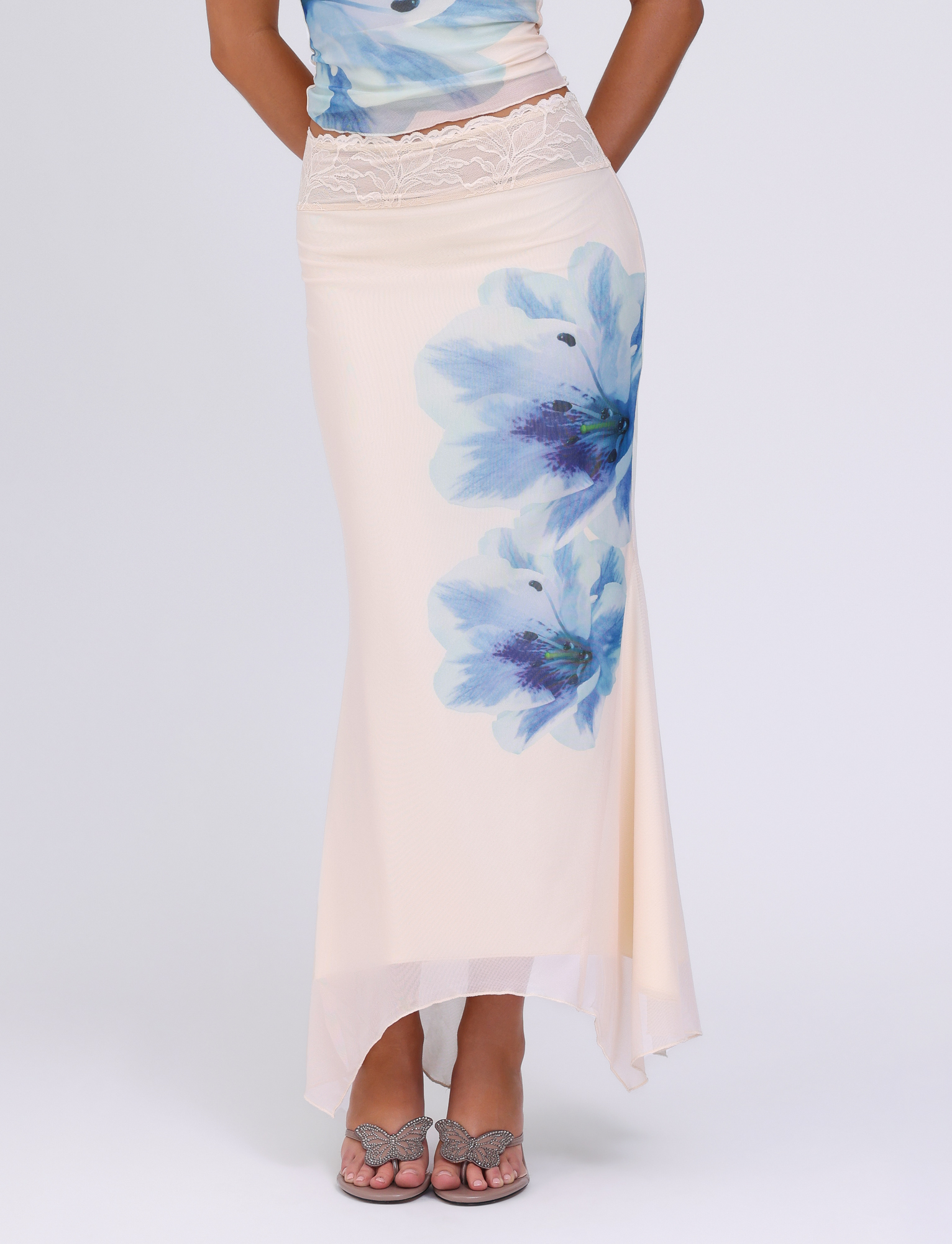 A person models the TIGERMIST LILLIAN SKIRT in white/cream with large blue floral print and lace waistband, paired with open-toe sandals, against a plain white background.