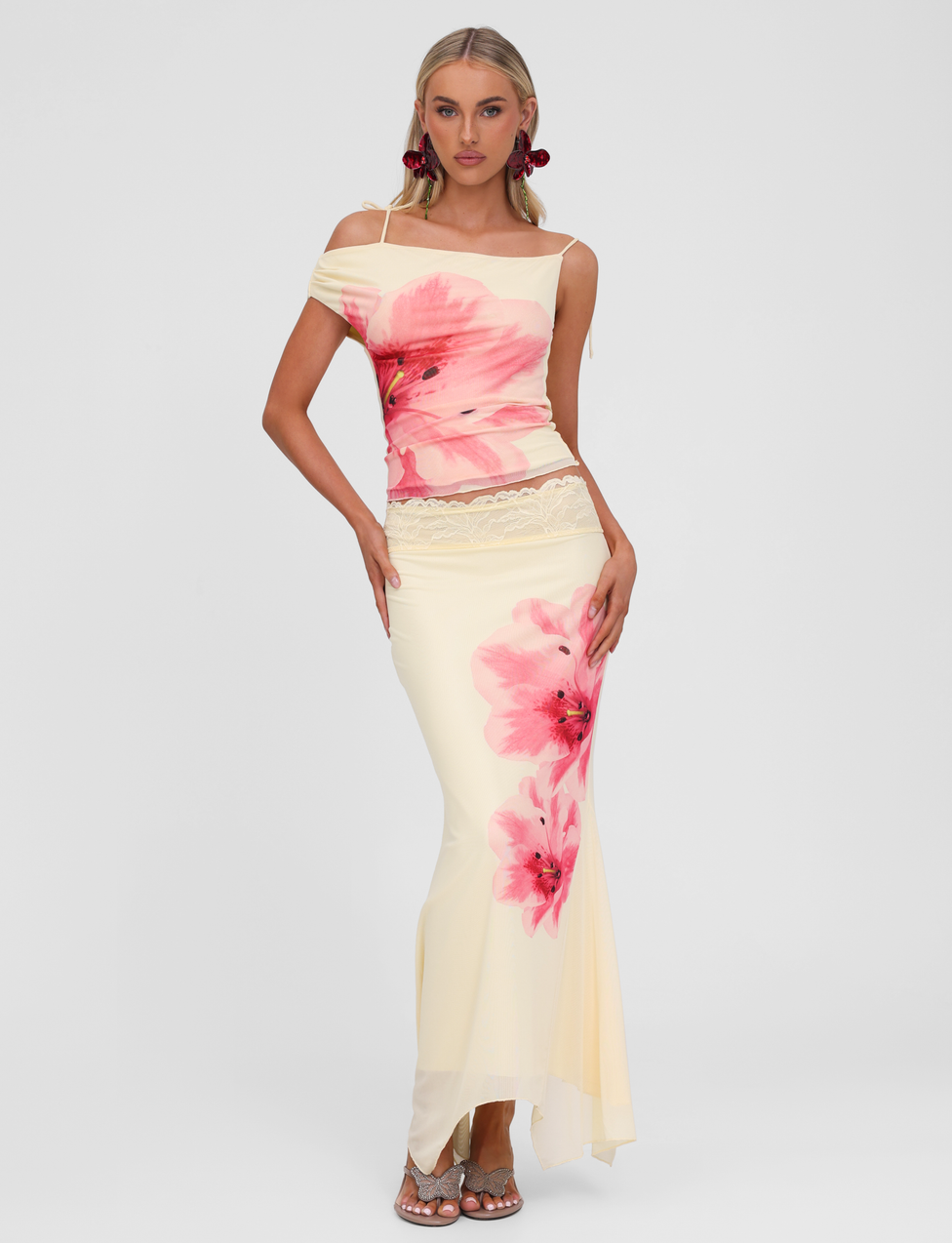 A woman models the TIGERMIST LILLIAN SKIRT in yellow, featuring large pink floral prints and a lace trim waistband. She pairs it with a cream off-shoulder top, silver heels, and pink statement earrings against a white background.