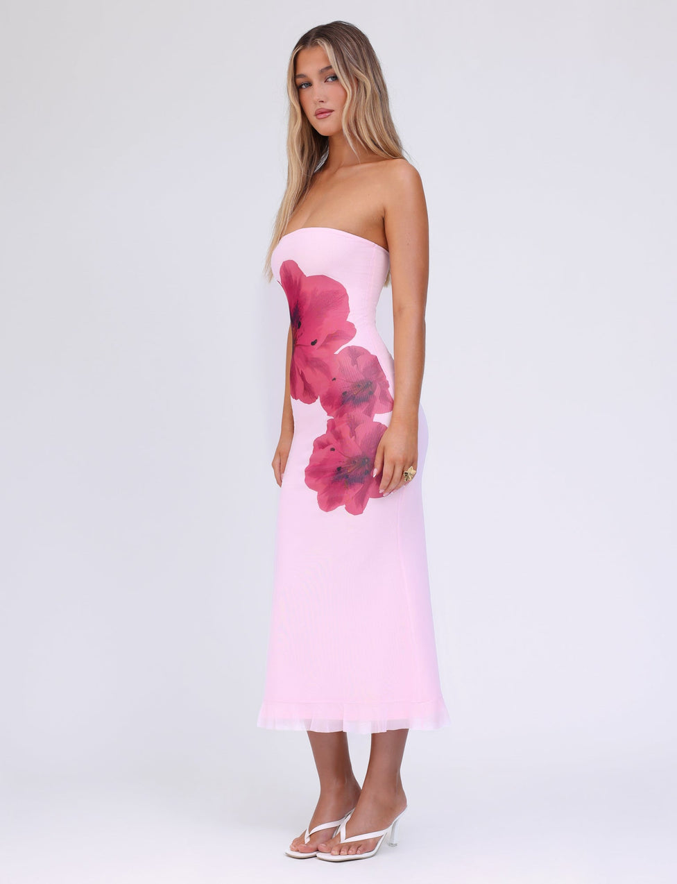 A woman stands wearing the strapless LILLIAN DRESS in baby pink from TIGERMIST, adorned with large red floral prints on the side. Her long, wavy hair cascades down as she pairs the midi-length dress with white, open-toe high heels. The background is plain white.