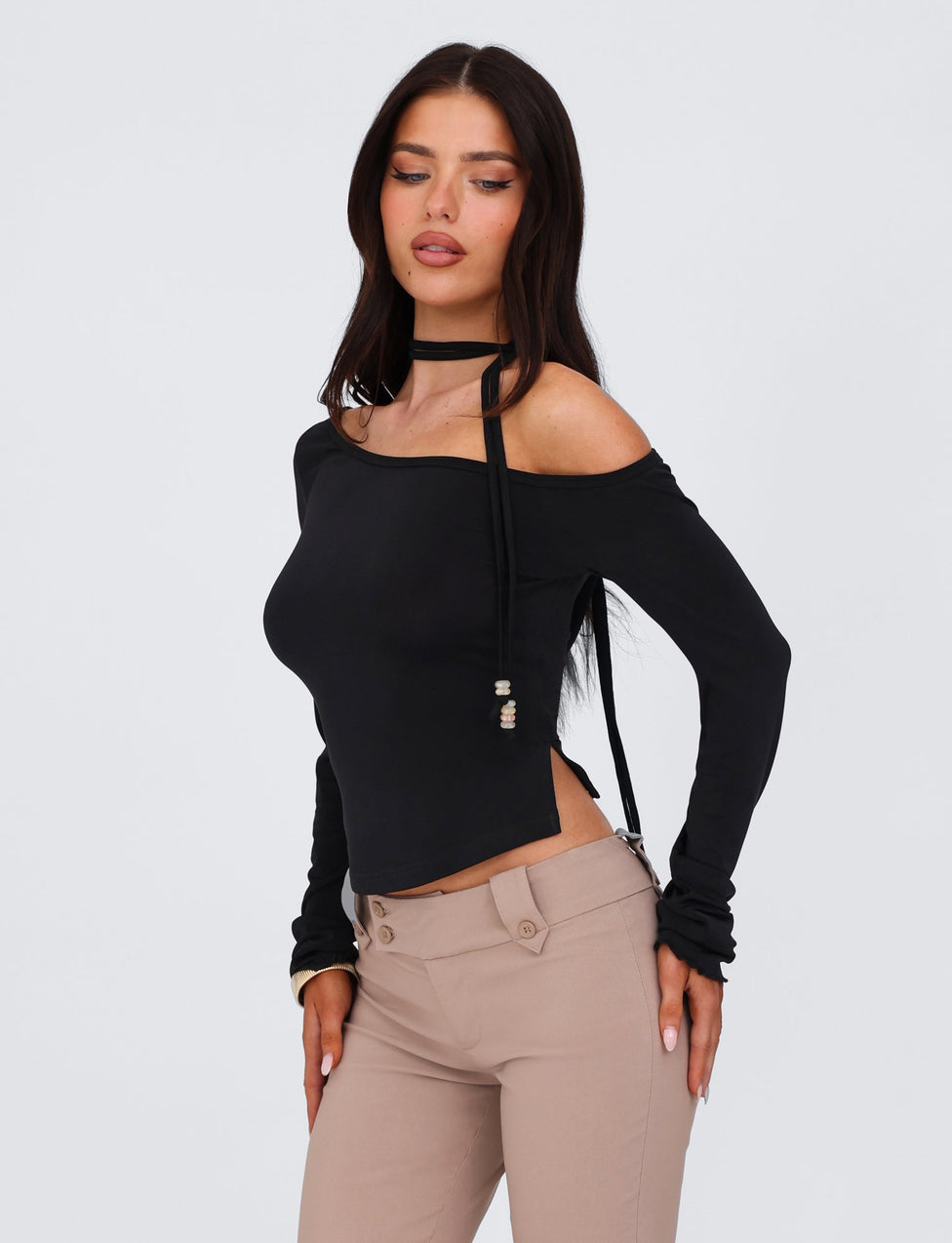 A woman with long dark hair is wearing an off-the-shoulder MIETTA TOP - BLACK by TIGERMIST, featuring a multi-tie neck, paired with taupe pants. She stands against a neutral background, looking slightly down and to the side, with her hands resting on her thighs.