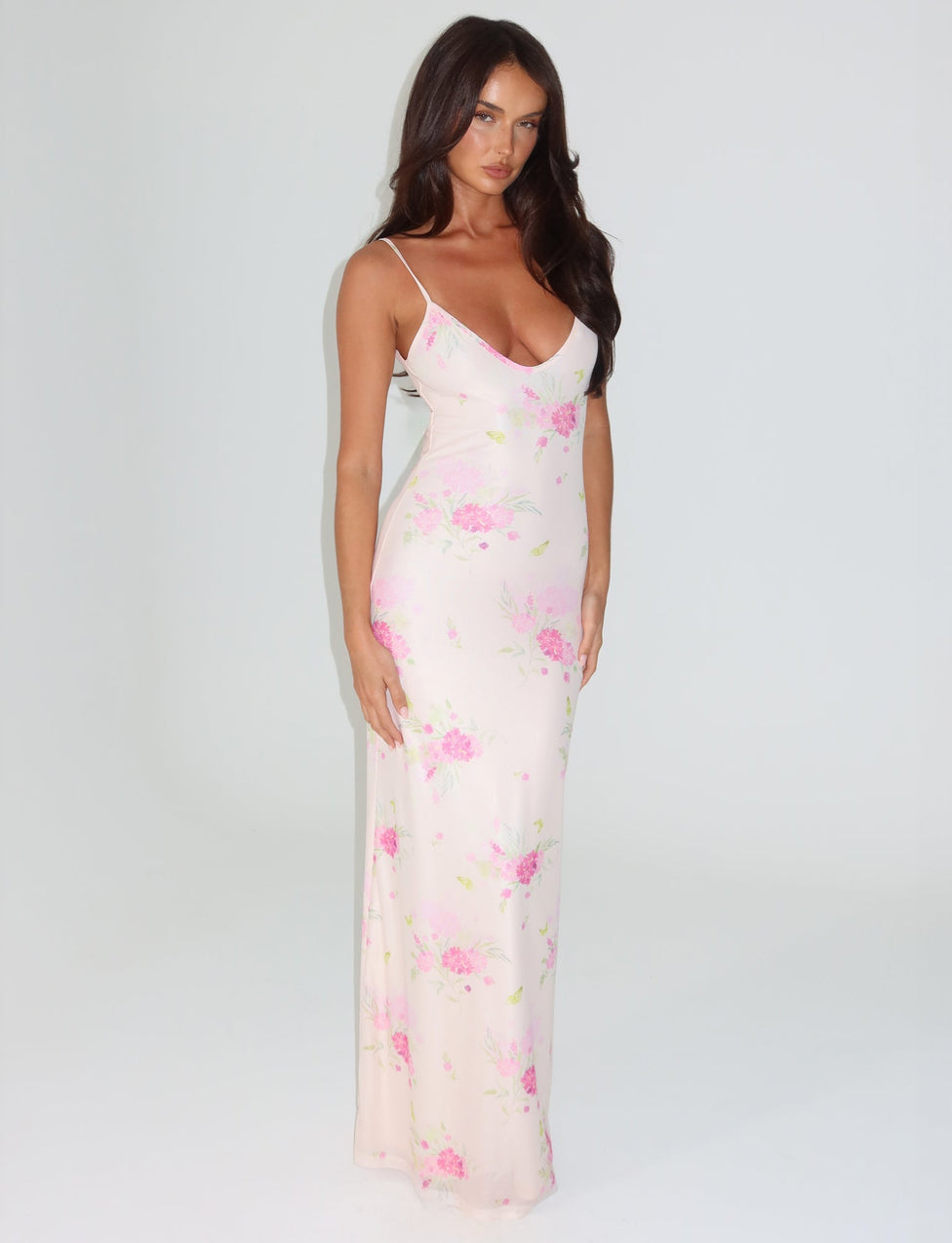 A woman in the OSIRIS DRESS by TIGERMIST stands against a plain white background. The maxi-length dress, featuring mesh fabrication and thin straps, showcases a beautiful watercolour floral pattern in pink. She has long, dark hair and is looking towards the camera.